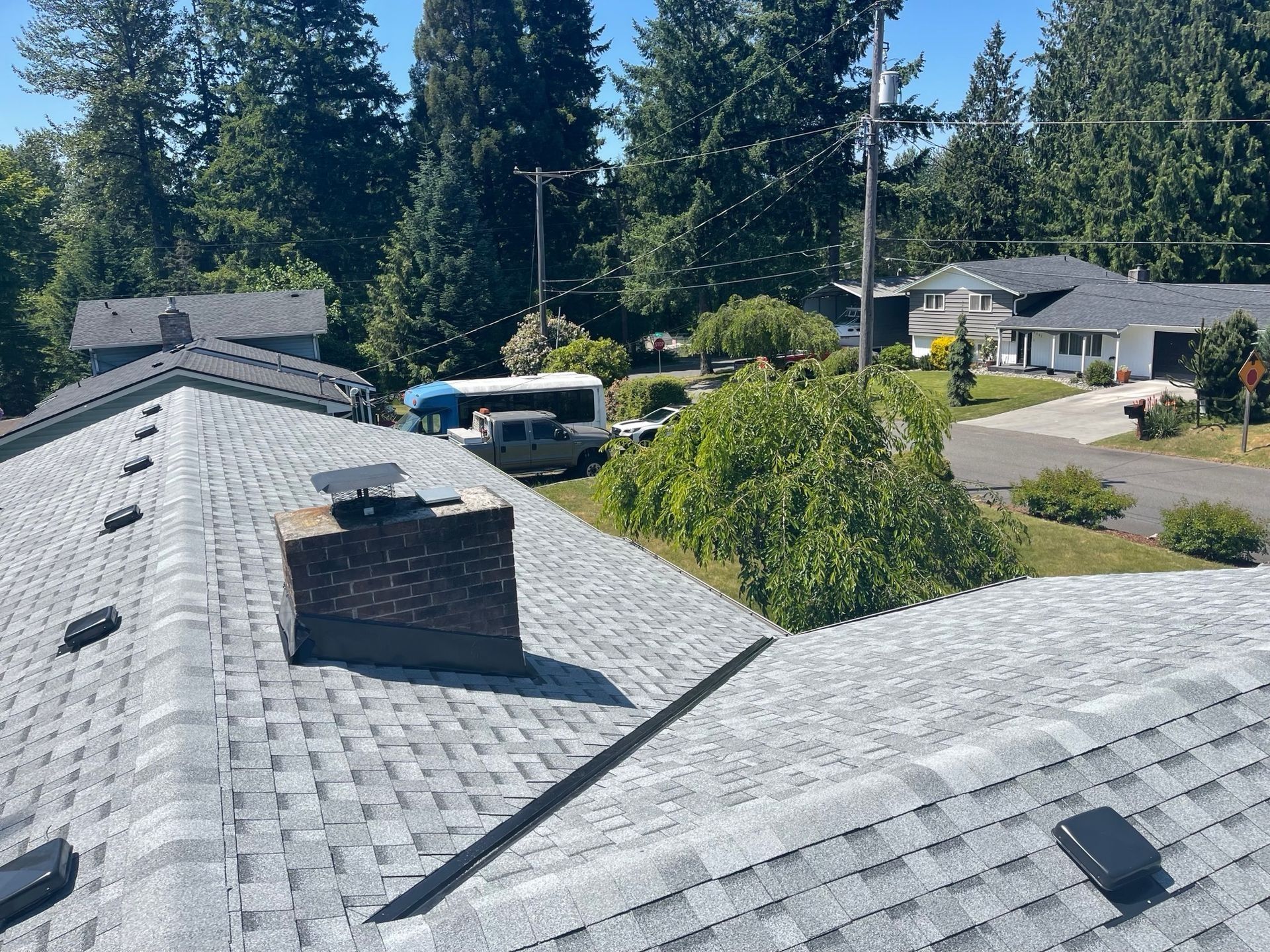 Gray asphalt shingle roof with a brick chimney on a sunny day. Houses and trees are in the background.