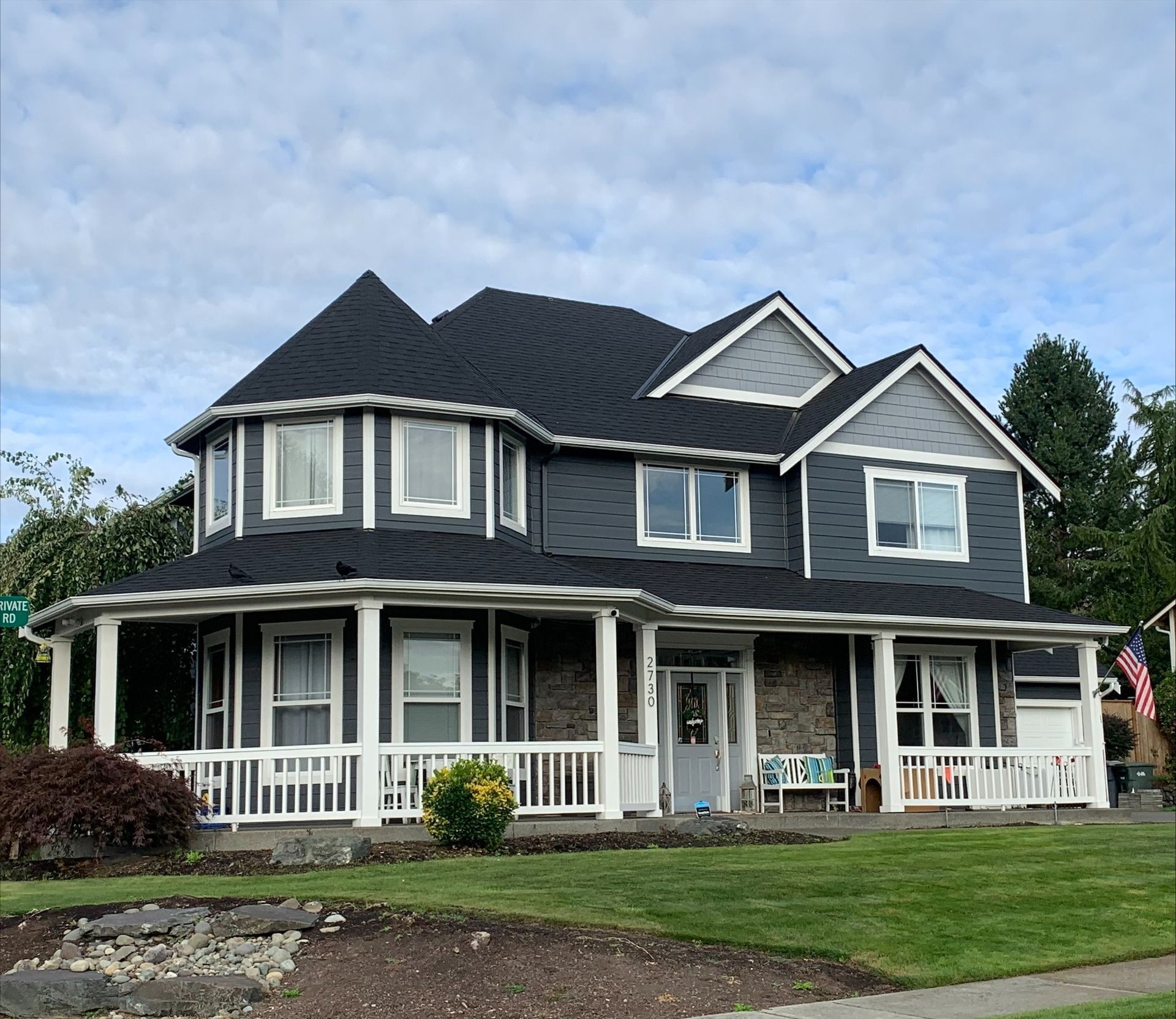 Two-story blue house with wrap-around porch, white trim, and a lawn under a blue sky.