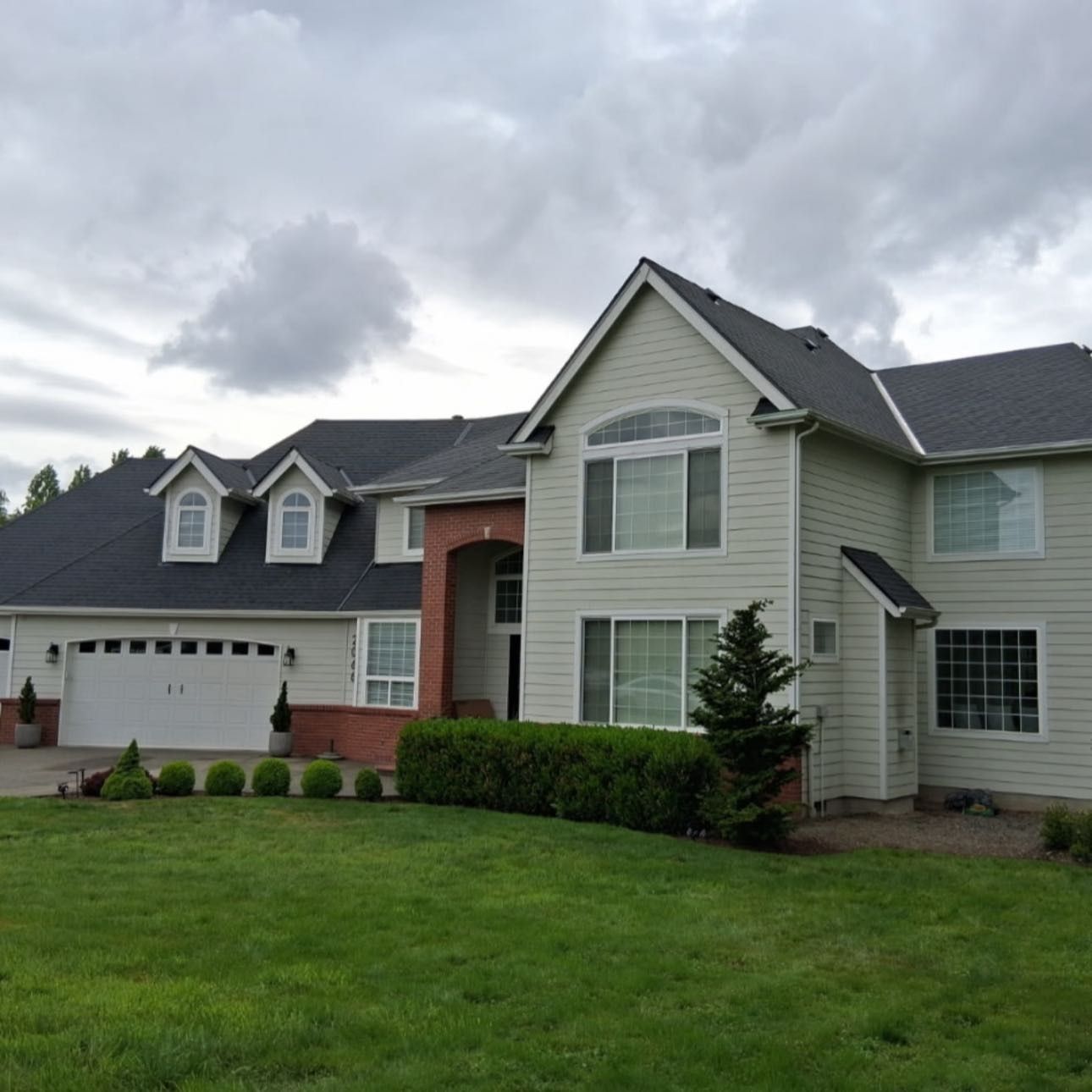 Two-story house with green siding, a black roof, a brick entrance, and a well-manicured lawn under a cloudy sky.