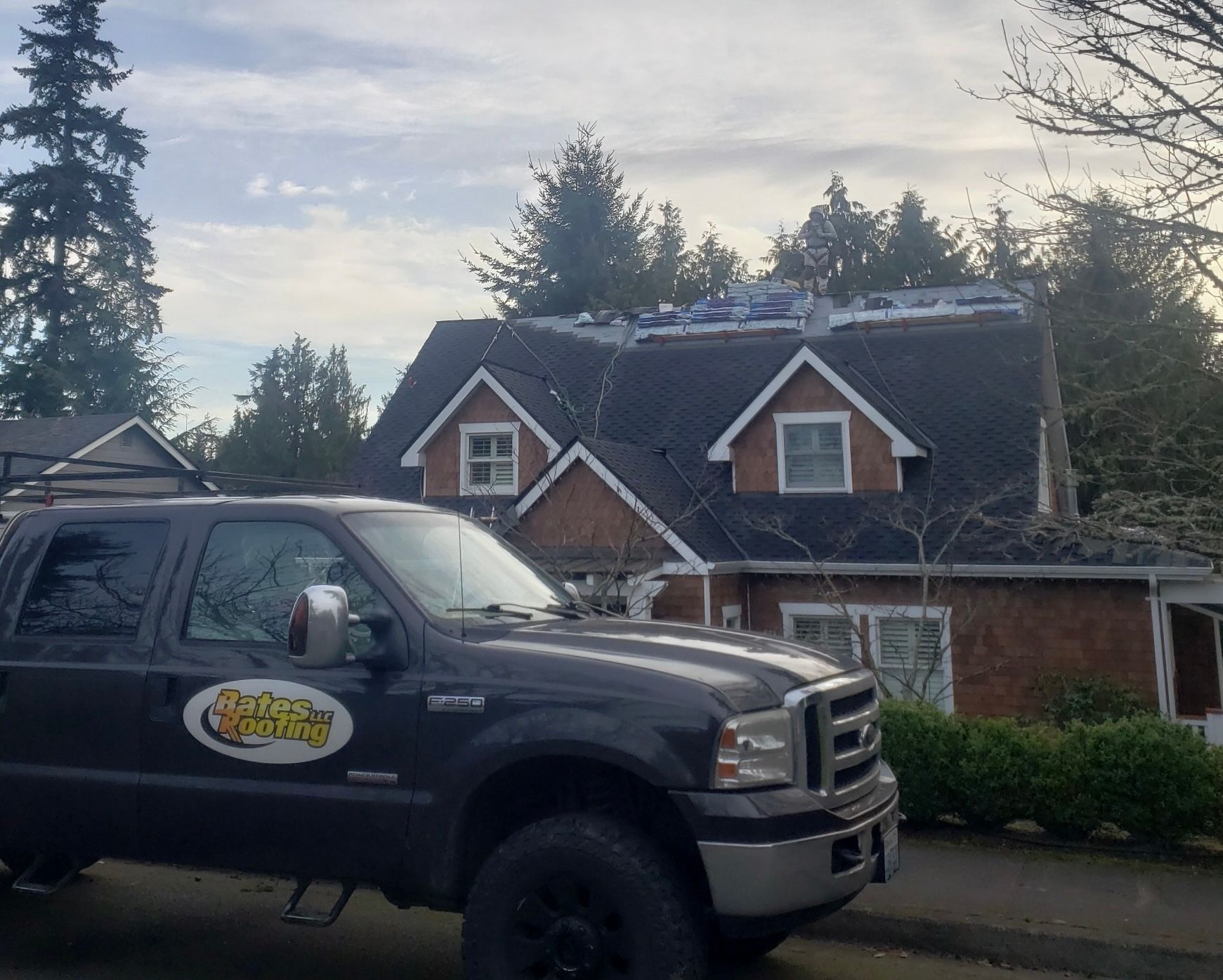 Dark truck in front of a house with partially removed roofing, trees in the background, overcast sky.