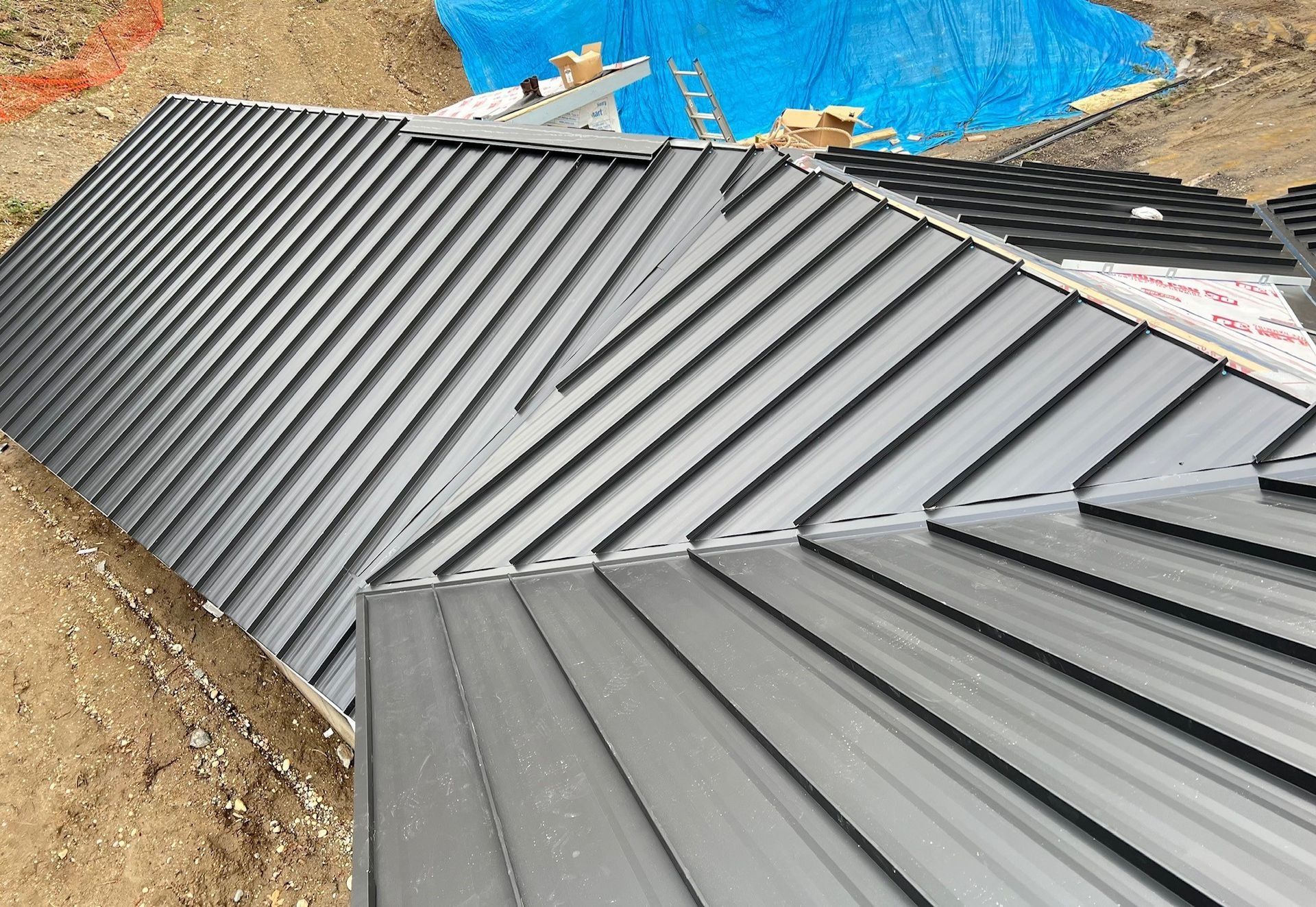 Metal roof installation in progress; angled, grey panels; dirt ground and blue tarp in the background.