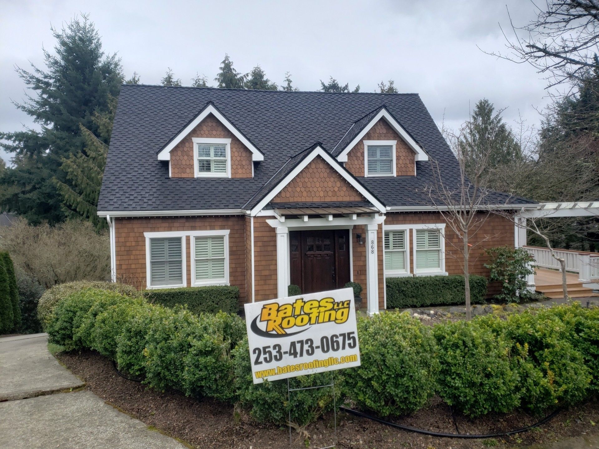 Small house with a dark roof and sign for Bates Roofing, bushes in front.