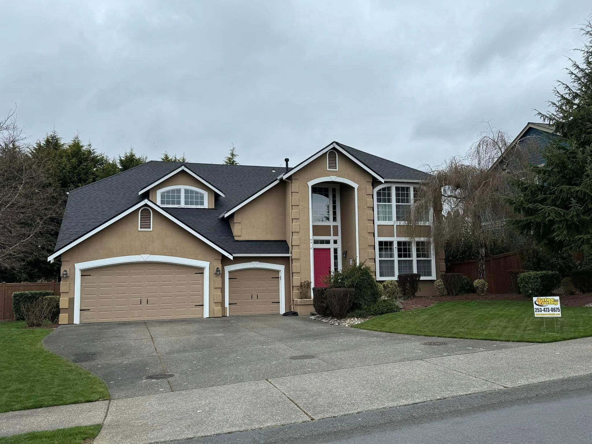Two-story house with tan stucco exterior, black roof, and red front door. Driveway and lawn in front. Overcast sky.