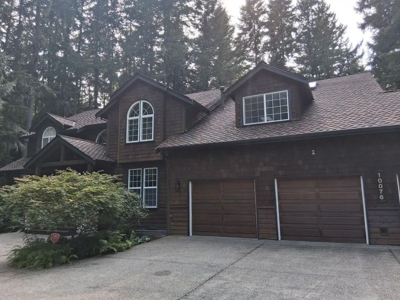 Brown house with two-car garage, multiple windows, and a tree-lined background.