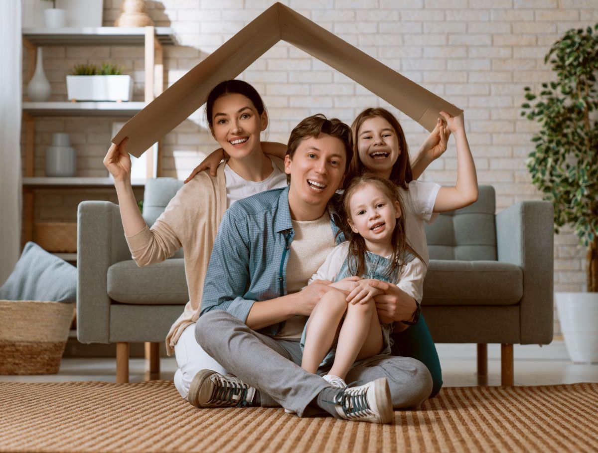 Family of four smiling, under cardboard house; sitting on rug in living room.