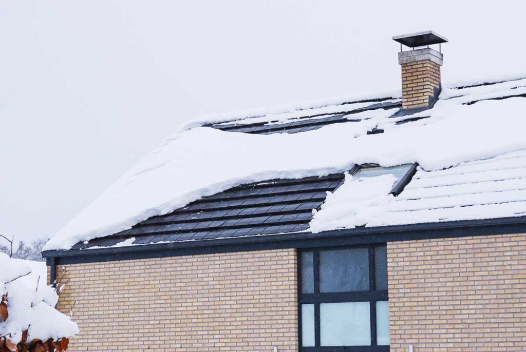 Snow-covered roof of a brick building with a chimney and a dark window, on a cloudy day.