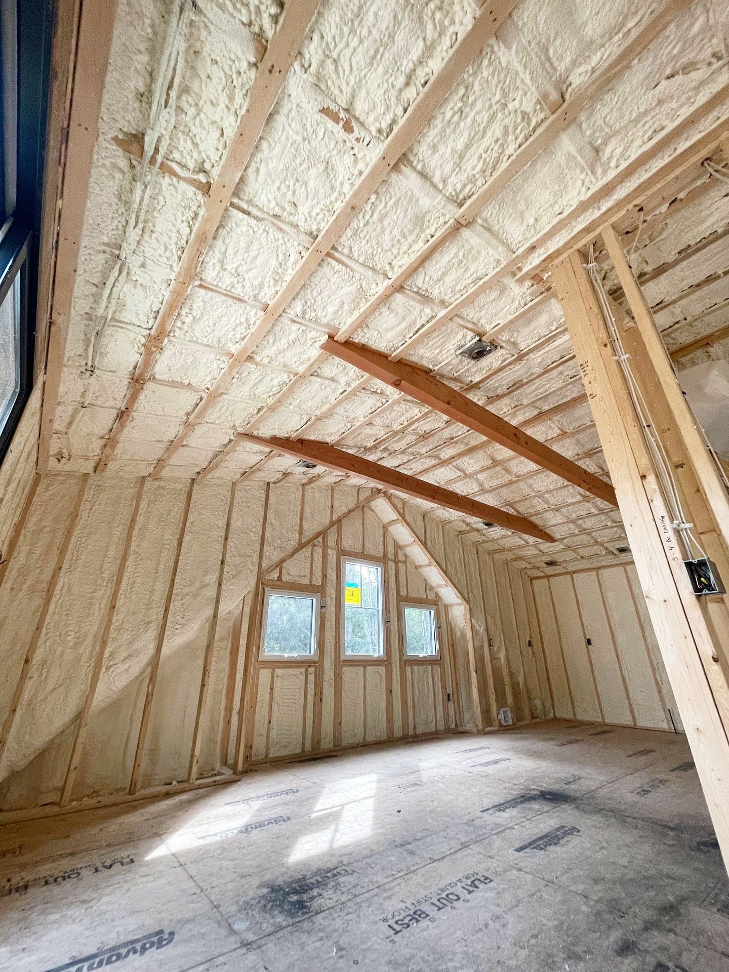 Interior view of an attic with spray foam insulation on the walls and ceiling. Wood beams, unfinished floor, and windows.