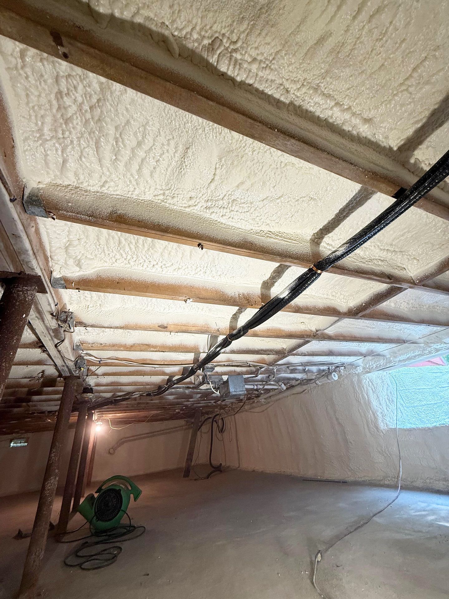 Basement with spray foam insulation on ceiling and walls, wooden beams, and concrete floor.