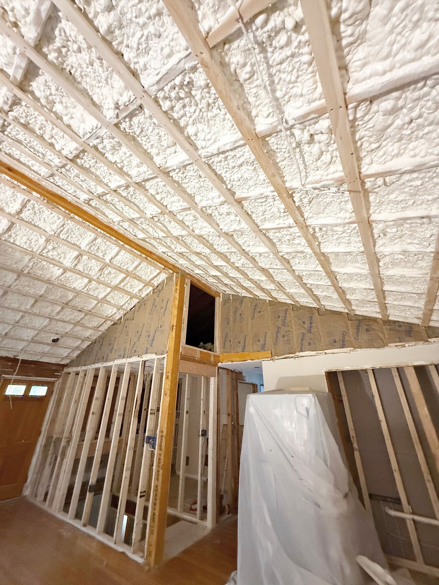 Interior view of a room under construction with spray foam insulation covering the ceiling. Wooden studs are visible.
