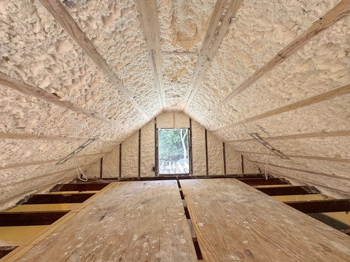 Interior attic space with spray foam insulation on rafters; wooden floor and window in the distance.