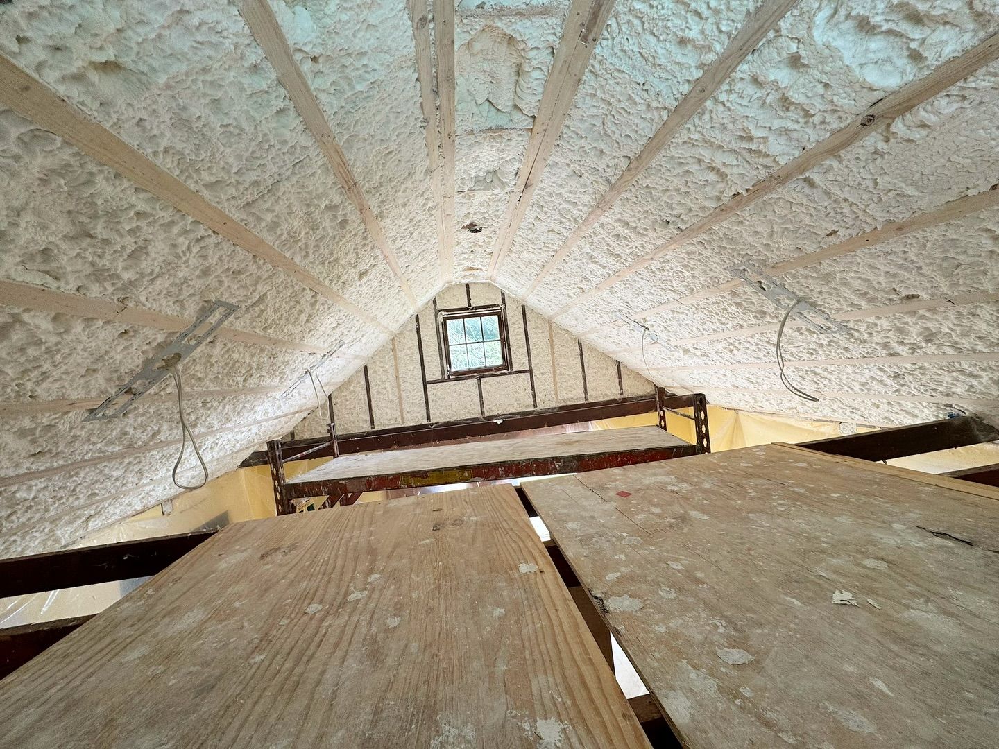 Attic interior with spray foam insulation on rafters and wood beams. A small window is visible.