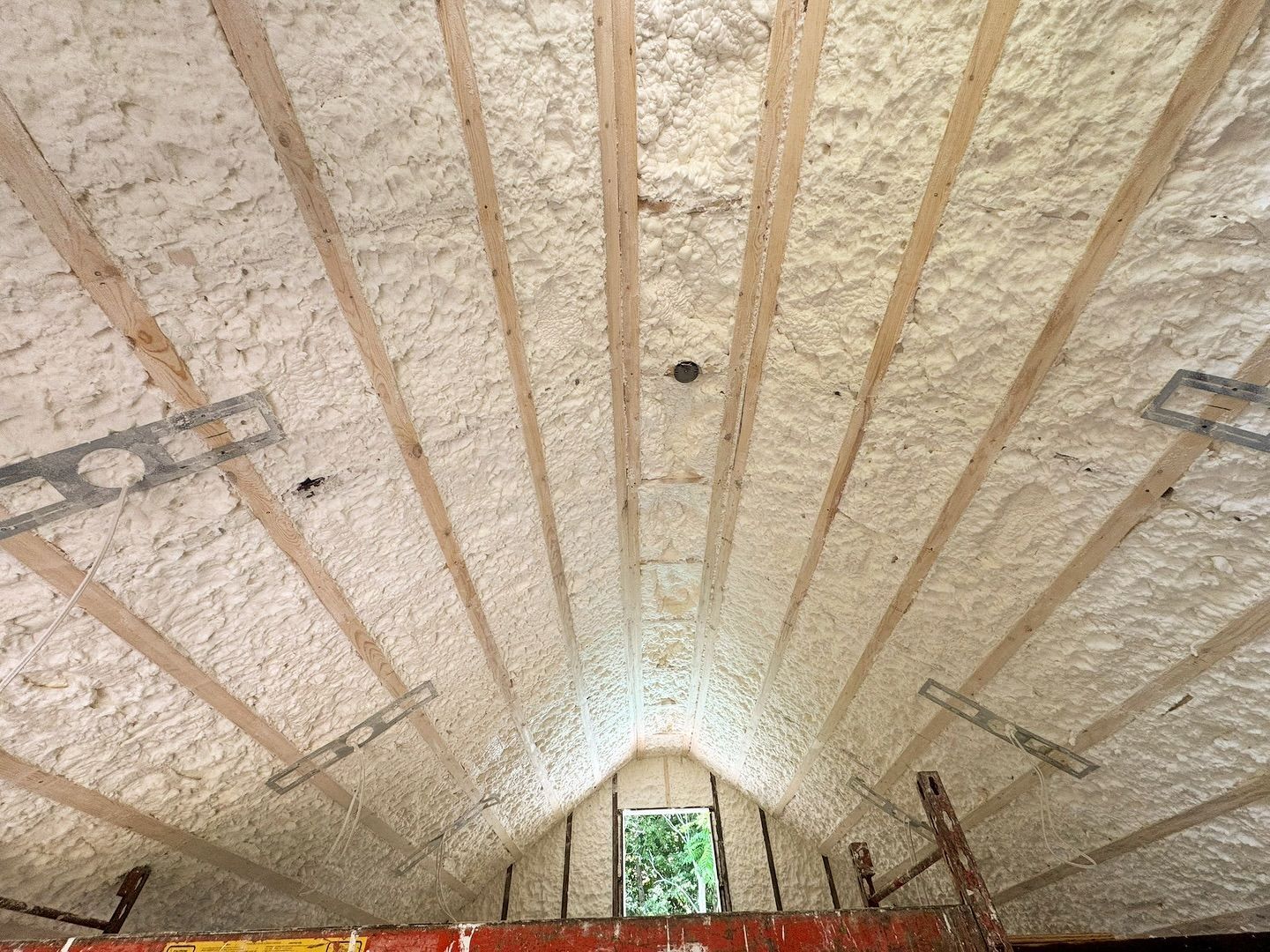 Attic ceiling with spray foam insulation between wooden beams, centered on a small window.