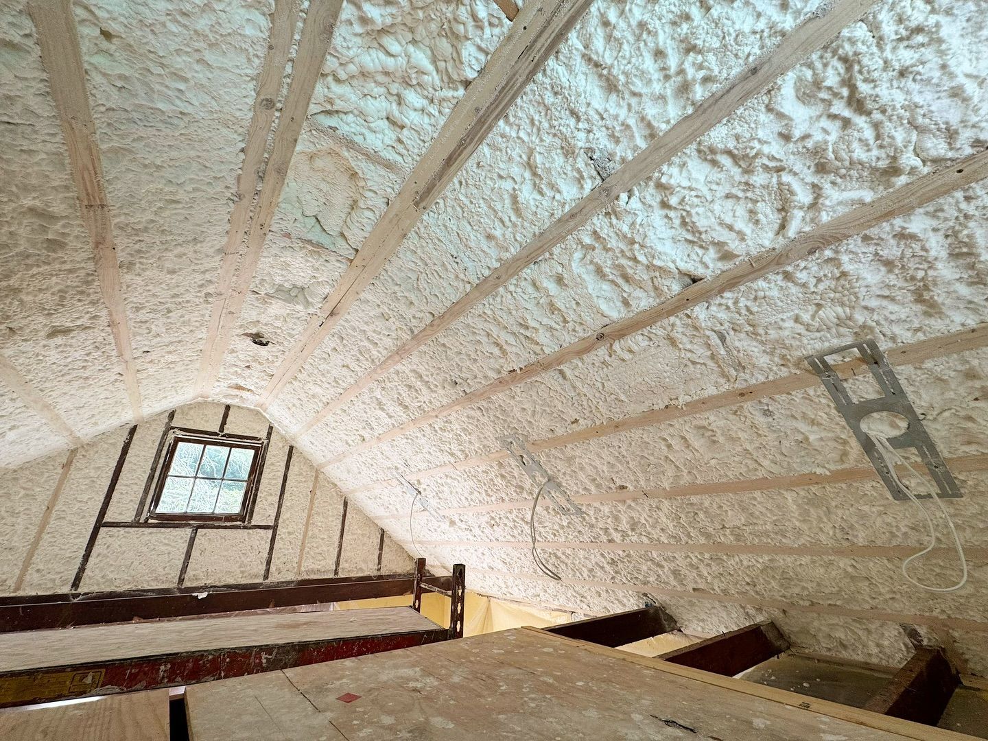 Attic interior insulated with light-colored spray foam, wooden beams and a small window visible.