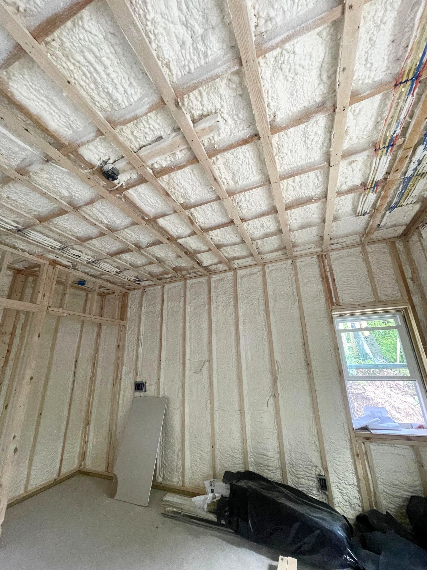Interior view of a room under construction, insulated with white spray foam on walls and ceiling. Wooden framing visible.