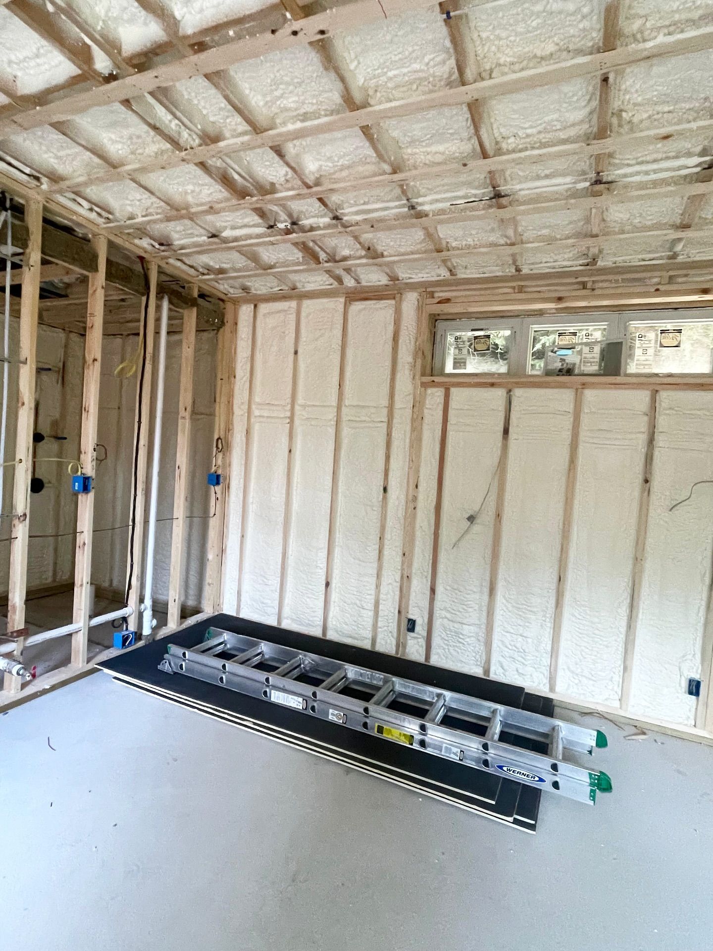 Interior of a room under construction with spray foam insulation on walls and ceiling. A ladder rests on a platform.