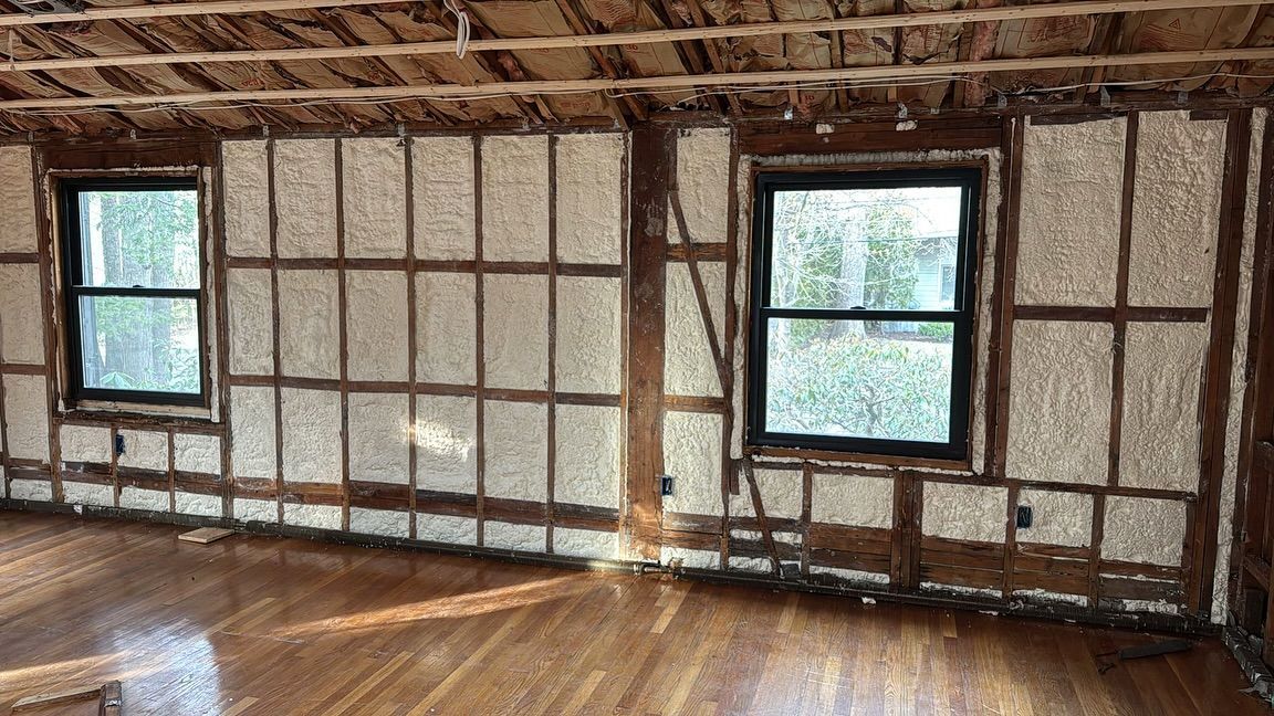 Interior room with foam insulation between wooden framing, two windows, and hardwood floors.