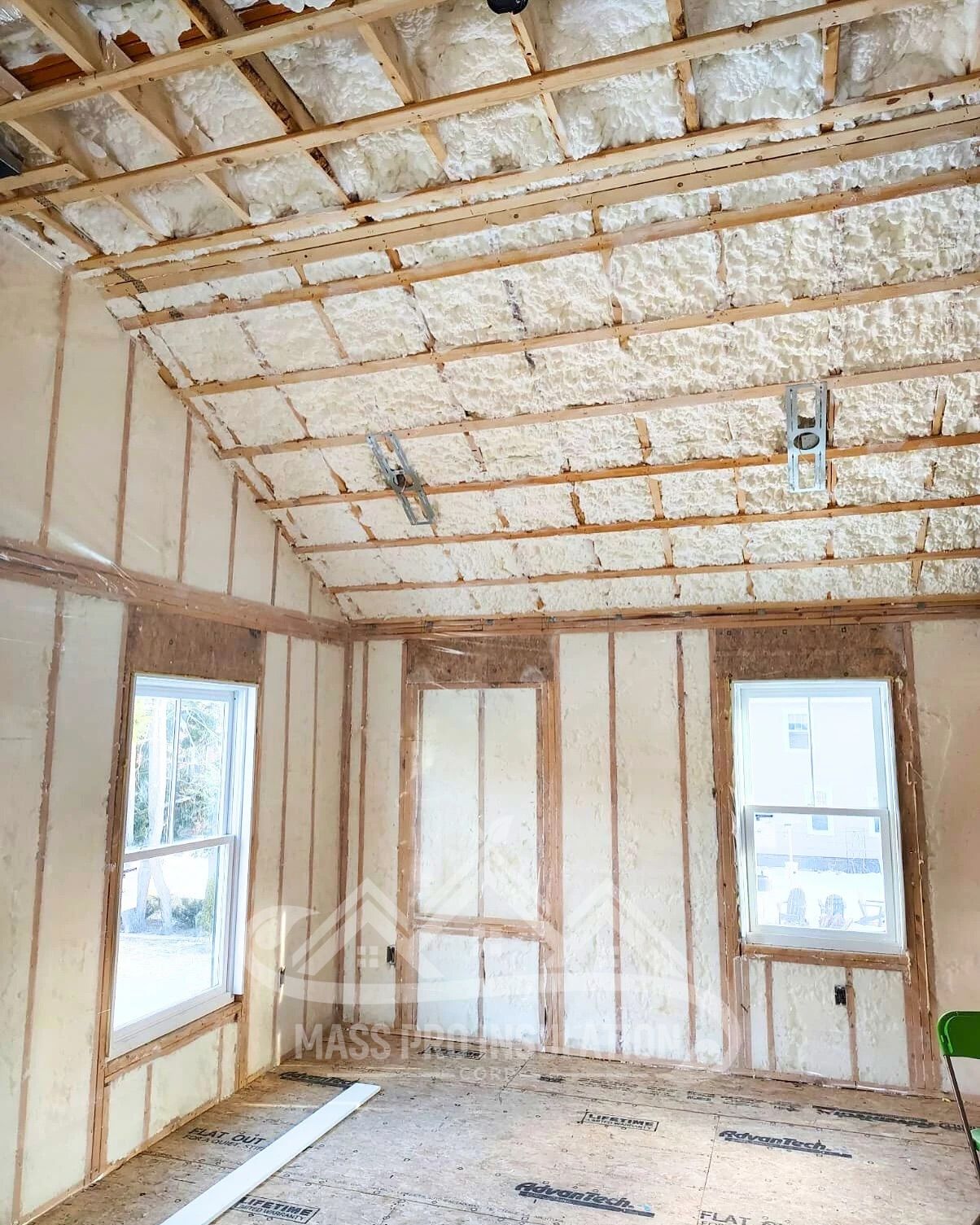 Interior view of a room under construction, with spray foam insulation covering walls and ceiling. Wooden framing is visible.