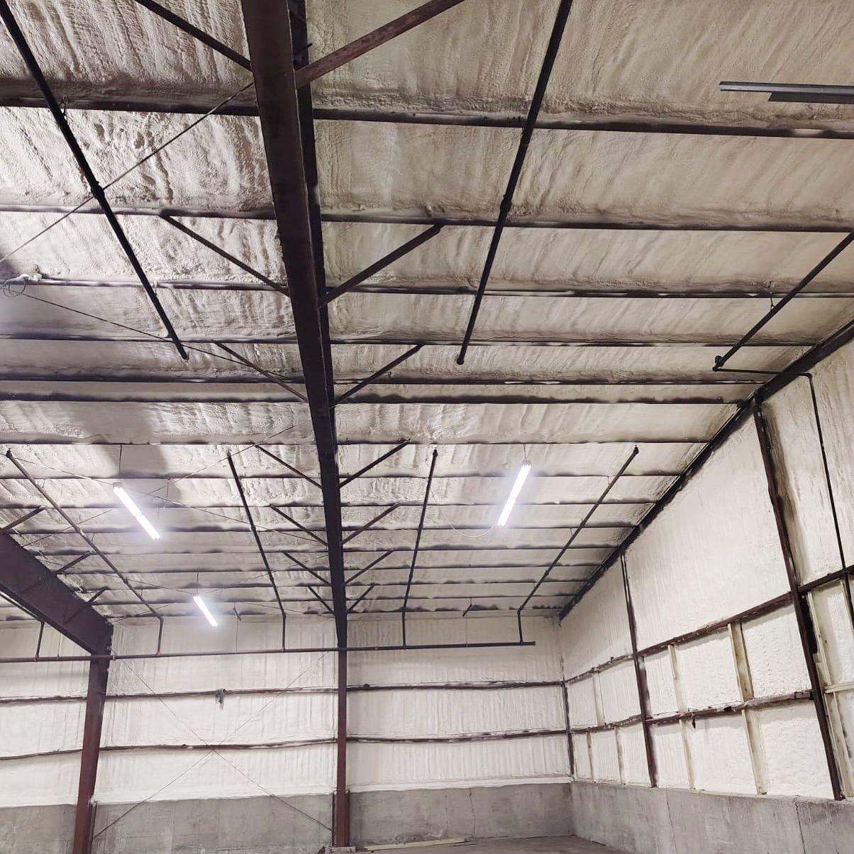 Interior view of a building with spray foam insulation on the walls and ceiling, metal beams, and fluorescent lights.