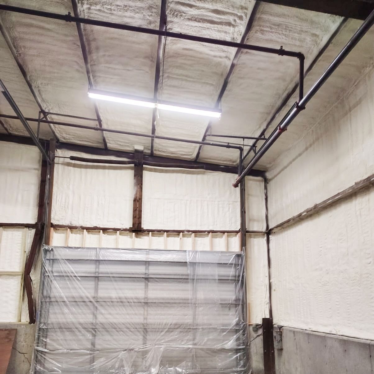 Warehouse interior insulated with white spray foam. Black metal beams and a closed garage door are visible.