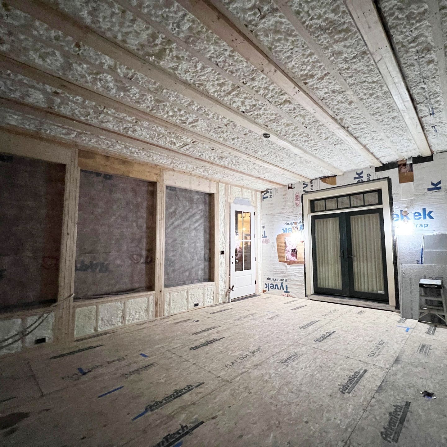 Interior of a room under construction: exposed beams, insulation, unfinished walls and floor, door and windows.