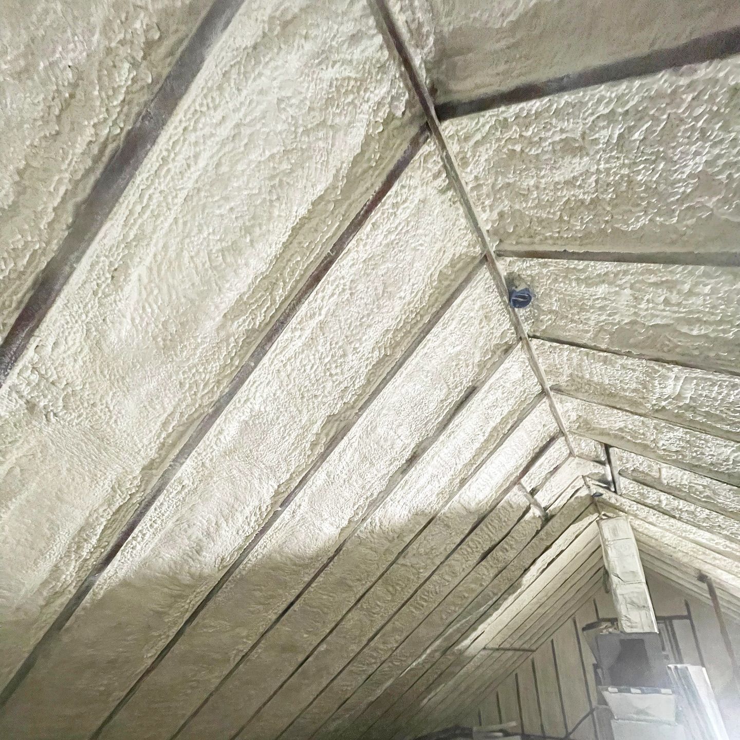 Spray foam insulation in an attic, covering rafters and creating a white, textured surface.