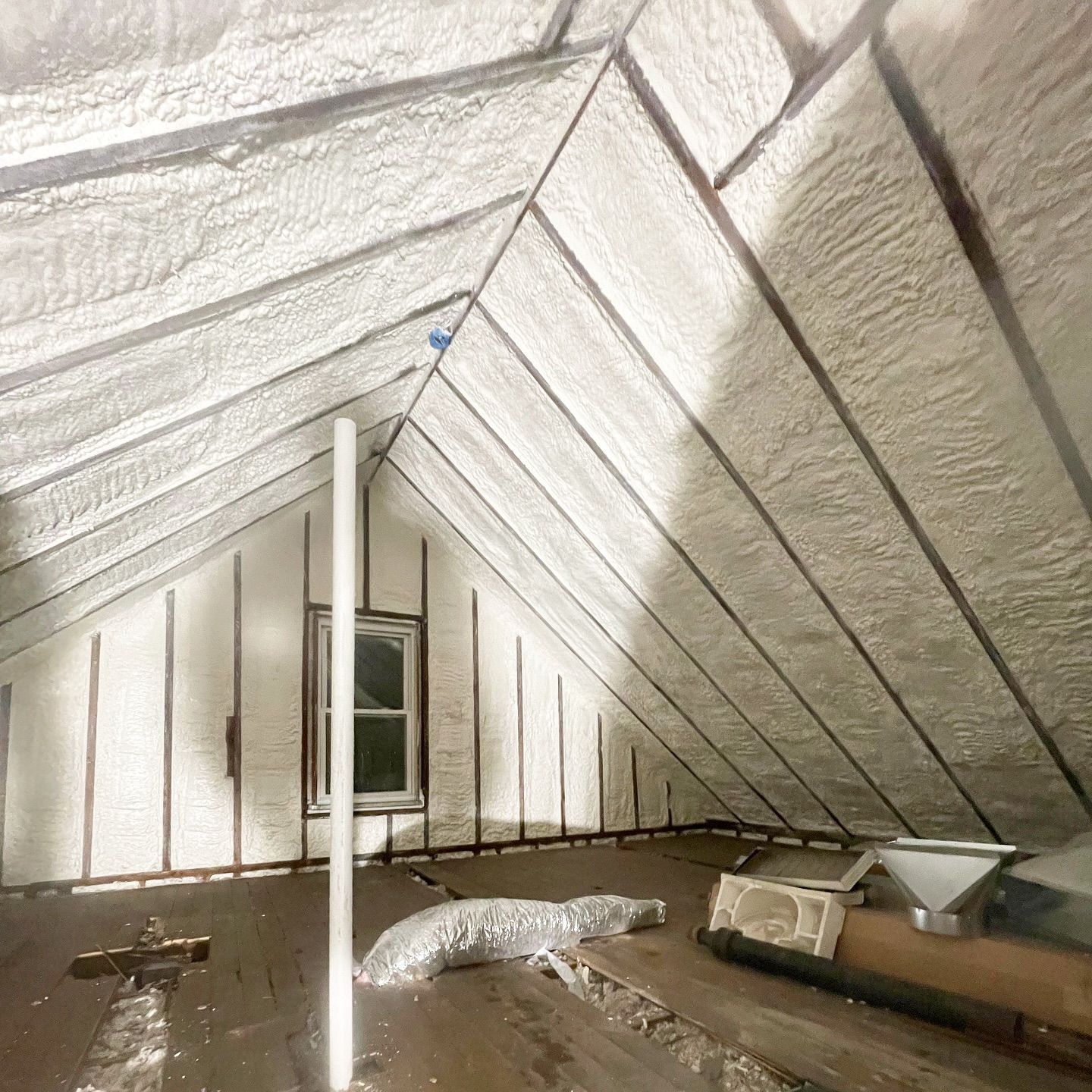 Attic with white spray foam insulation on rafters and walls, small window, vents, and exposed wood flooring.