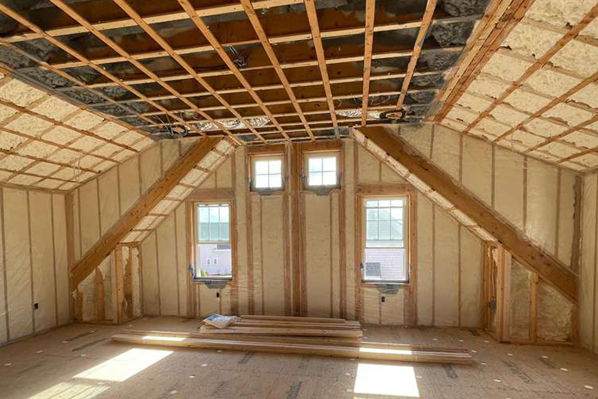 Interior of a room with spray foam insulation on walls and ceiling, unfinished windows and wood beams.