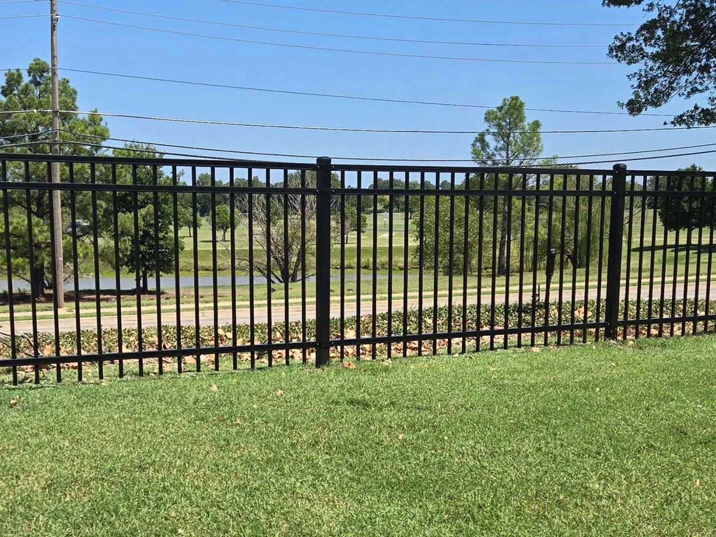Black metal fence on green grass, trees and water visible in the background under a blue sky.