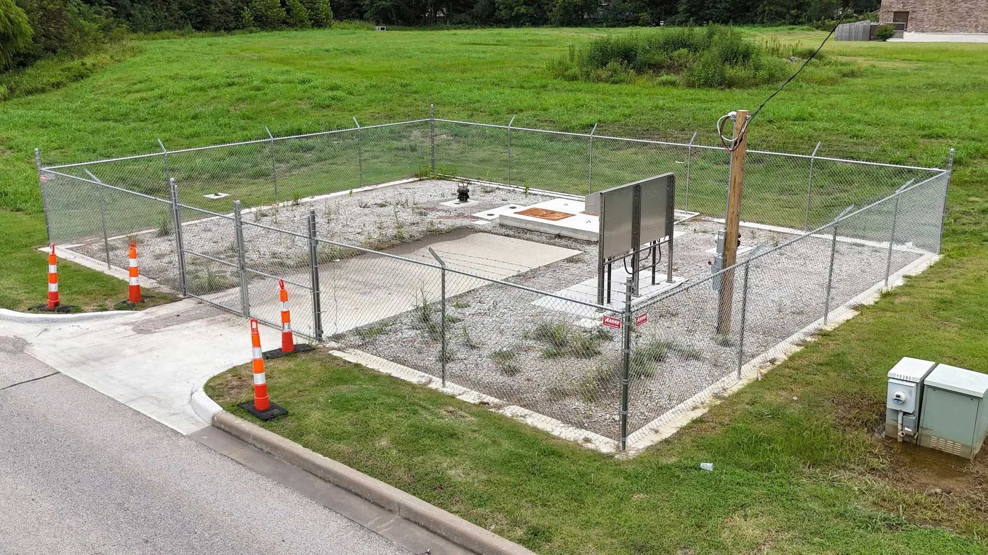 Fenced electrical equipment enclosure on concrete base, with power pole, surrounded by grass and a road.