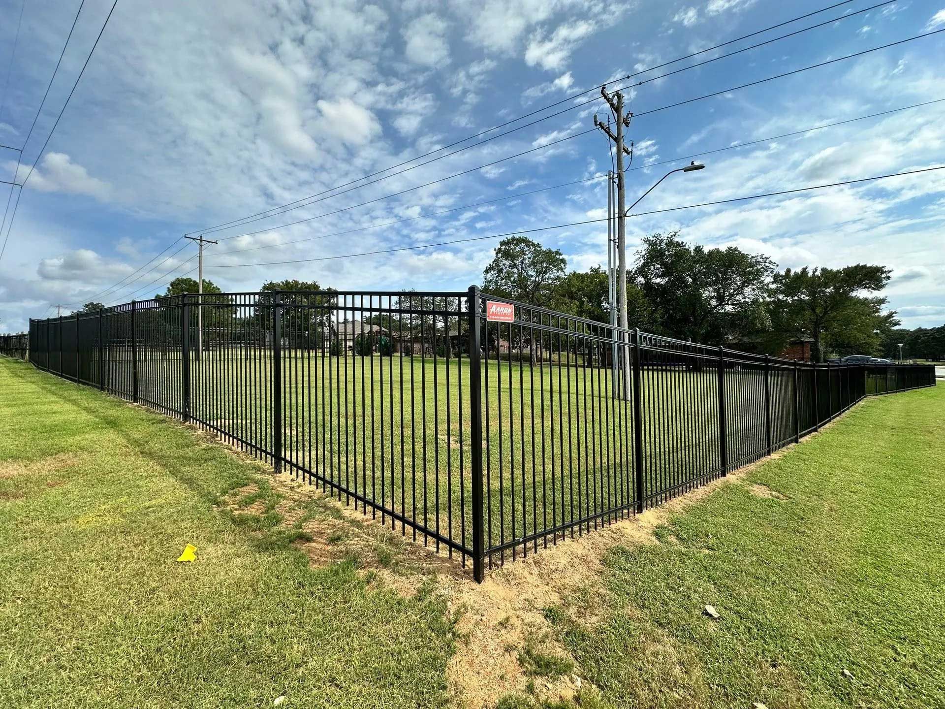Black metal fence surrounding a grassy area under a partly cloudy sky. Power lines and utility pole visible.