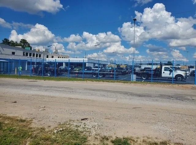 A fenced-in lot with vehicles parked behind a blue metal fence under a partly cloudy sky.