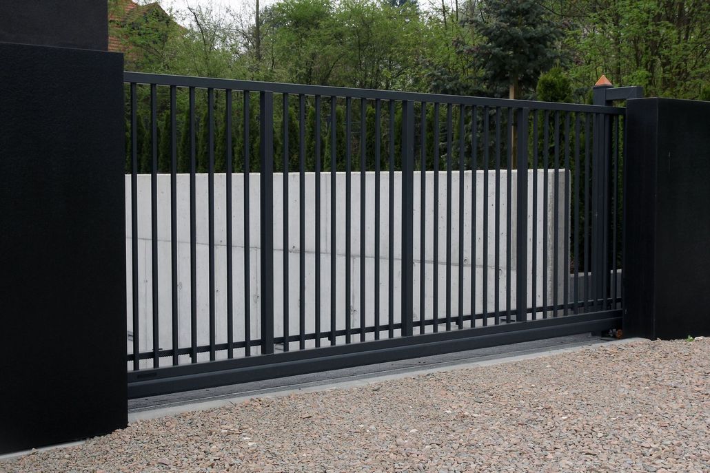 Black wrought-iron gate and fence in front of a driveway, with evergreen trees in the background.