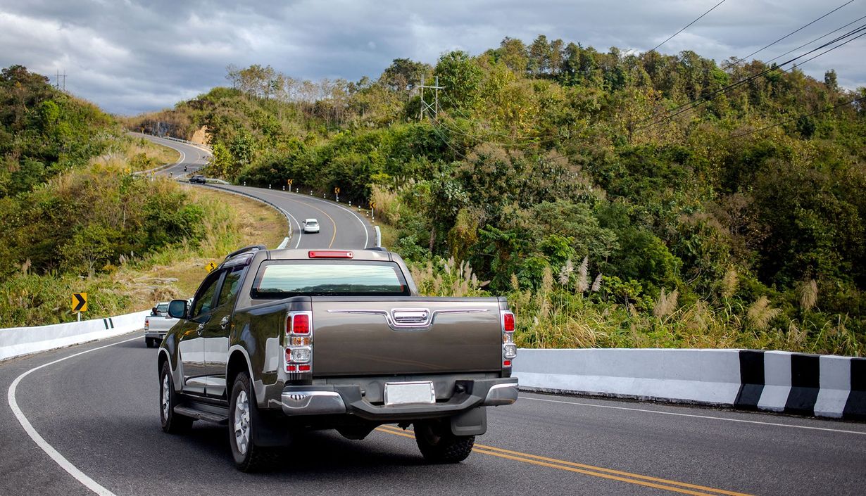 Pickup truck driving on a winding mountain road with a black-and-white barrier and green hillside.
