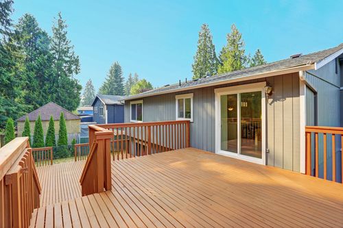 Wooden deck outside a gray house with sliding glass doors and trees in the background