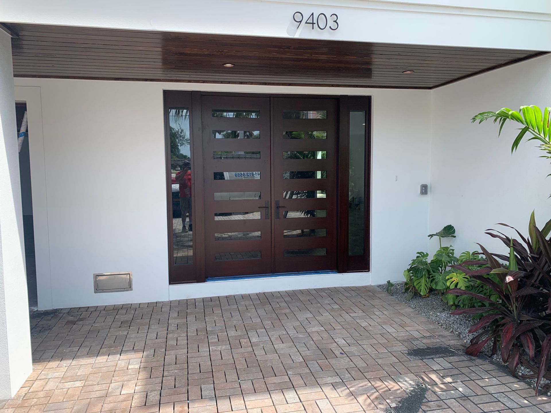 Front entrance with dark wooden double doors, white walls, brick walkway, and house number 9403 above the porch.