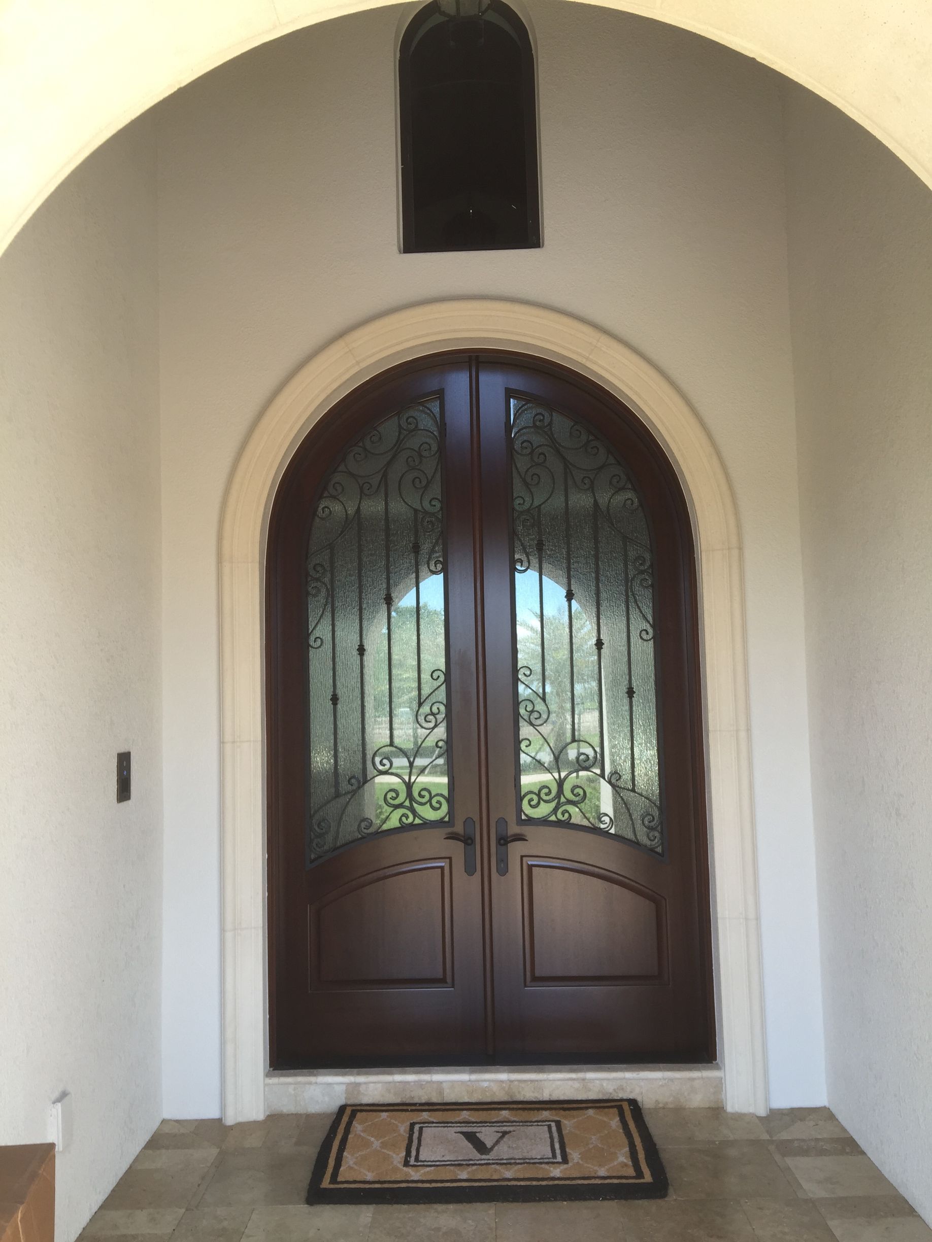 Arched entryway with dark wooden double doors, decorative glass, and a small welcome mat.