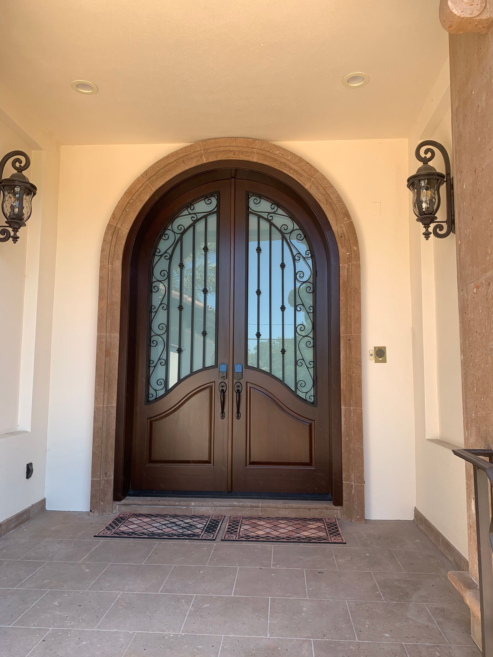 Front entrance with arched wooden double doors, frosted glass panels, and wall lanterns on a covered porch.