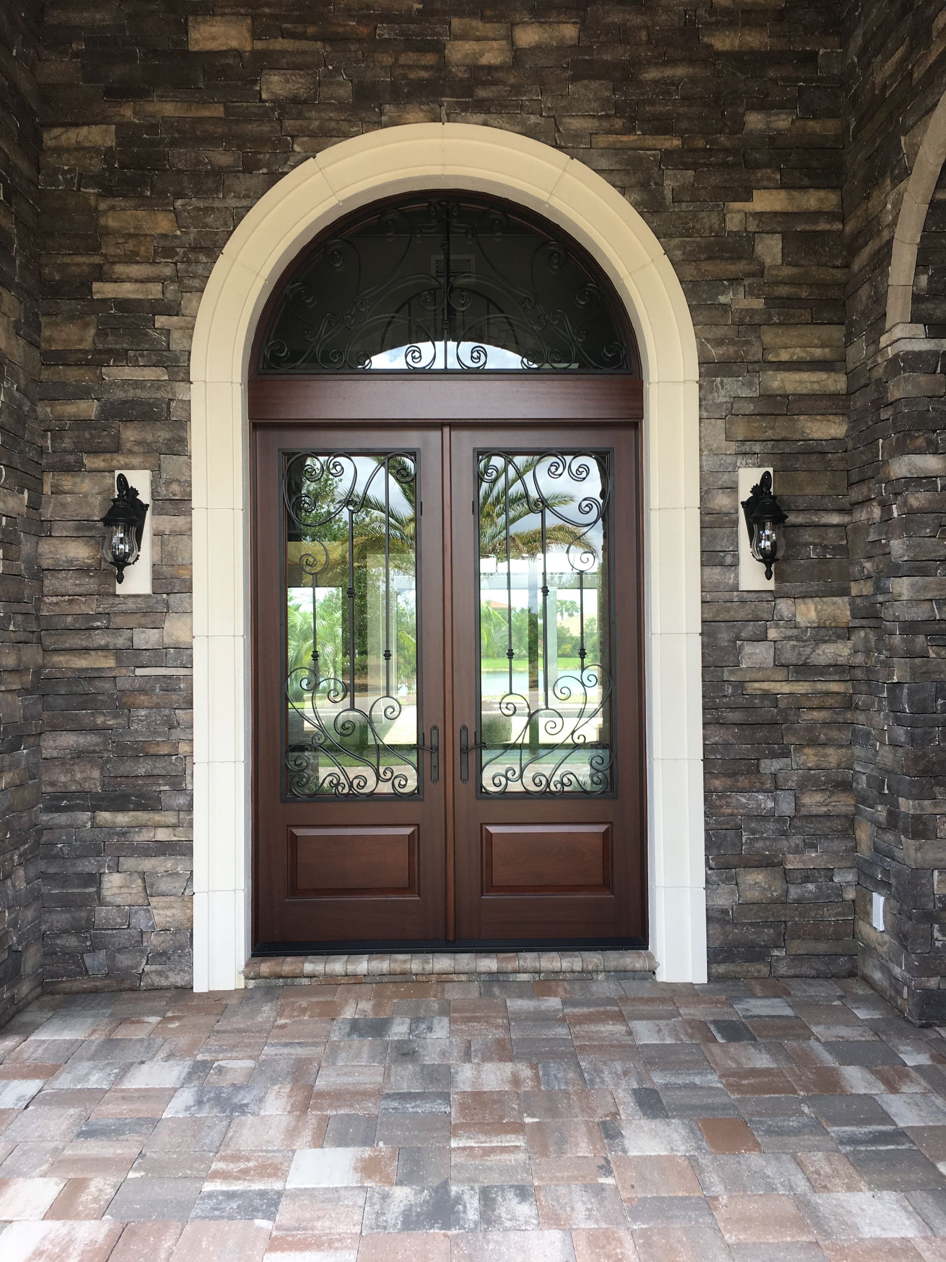 Arched double wooden doors in a stone entryway with brick flooring and lanterns on both sides