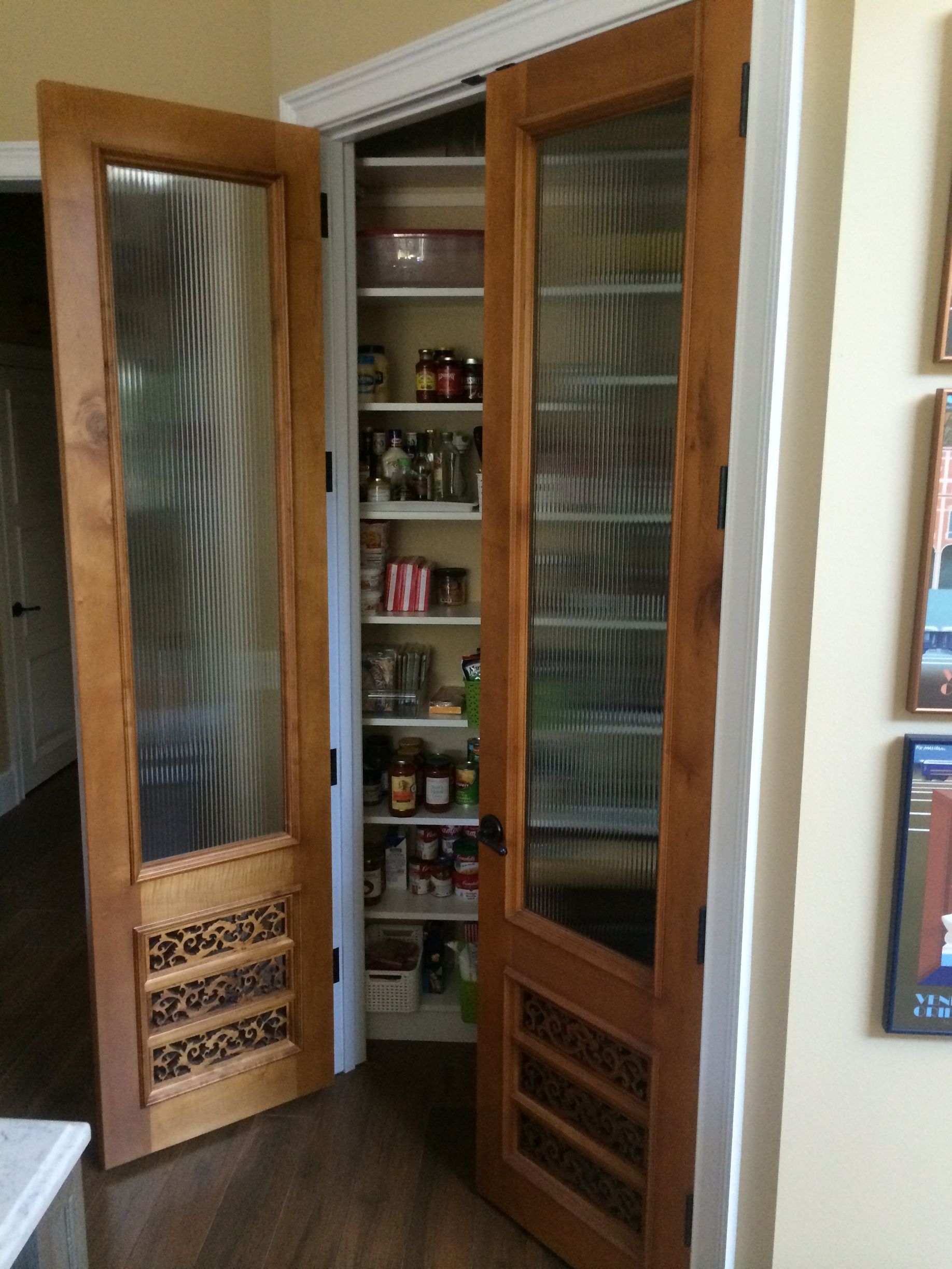 Open wooden pantry cabinet with glass doors, revealing shelves of bottles and canned goods in a kitchen.