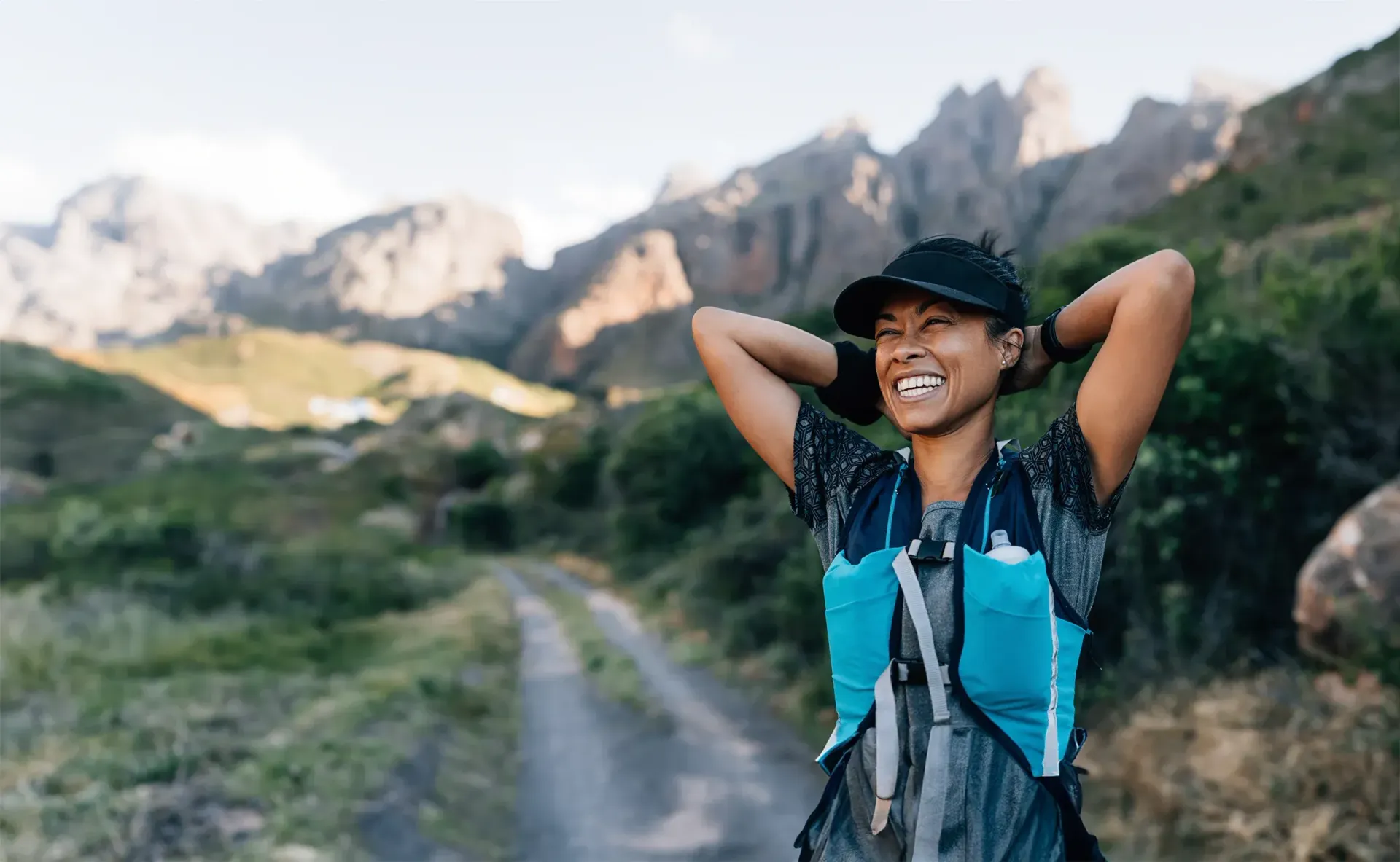 Woman smiles with arms behind head on a mountain trail, wearing a hat and hydration pack; rocky peaks in the background.