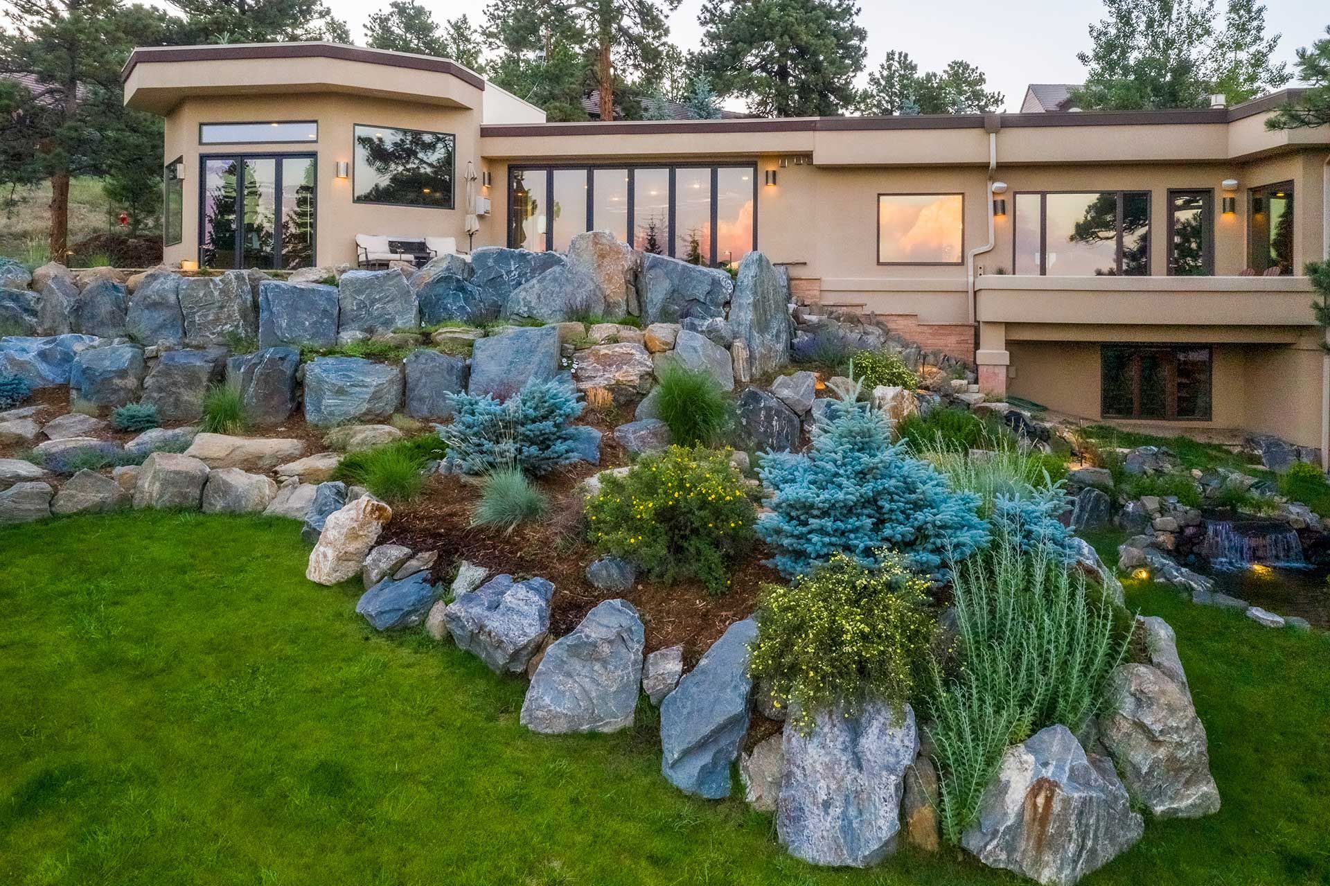 A modern multi-level house with a large terraced rock garden, evergreen shrubs, and a lush green lawn at dusk.