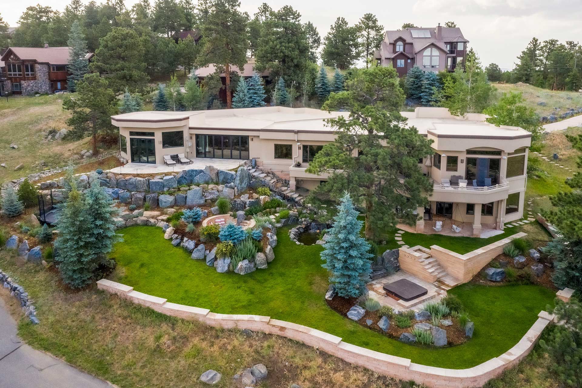 An elevated view of a modern, multi-level beige home with tiered stone landscaping, green lawns, and pine trees.