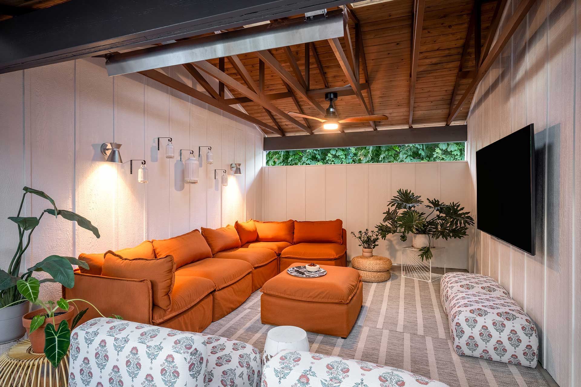Cozy patio lounge with an orange sectional, ottoman, patterned floor cushions, plants, and a wall-mounted television.