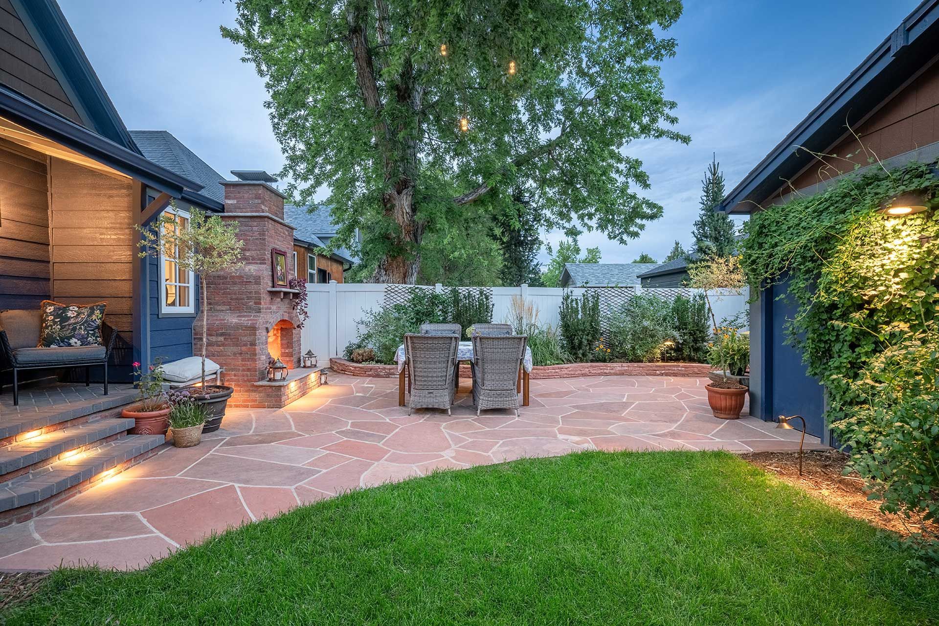 A backyard patio with a stone fireplace, outdoor seating, and a dining set, viewed from the lawn at dusk.