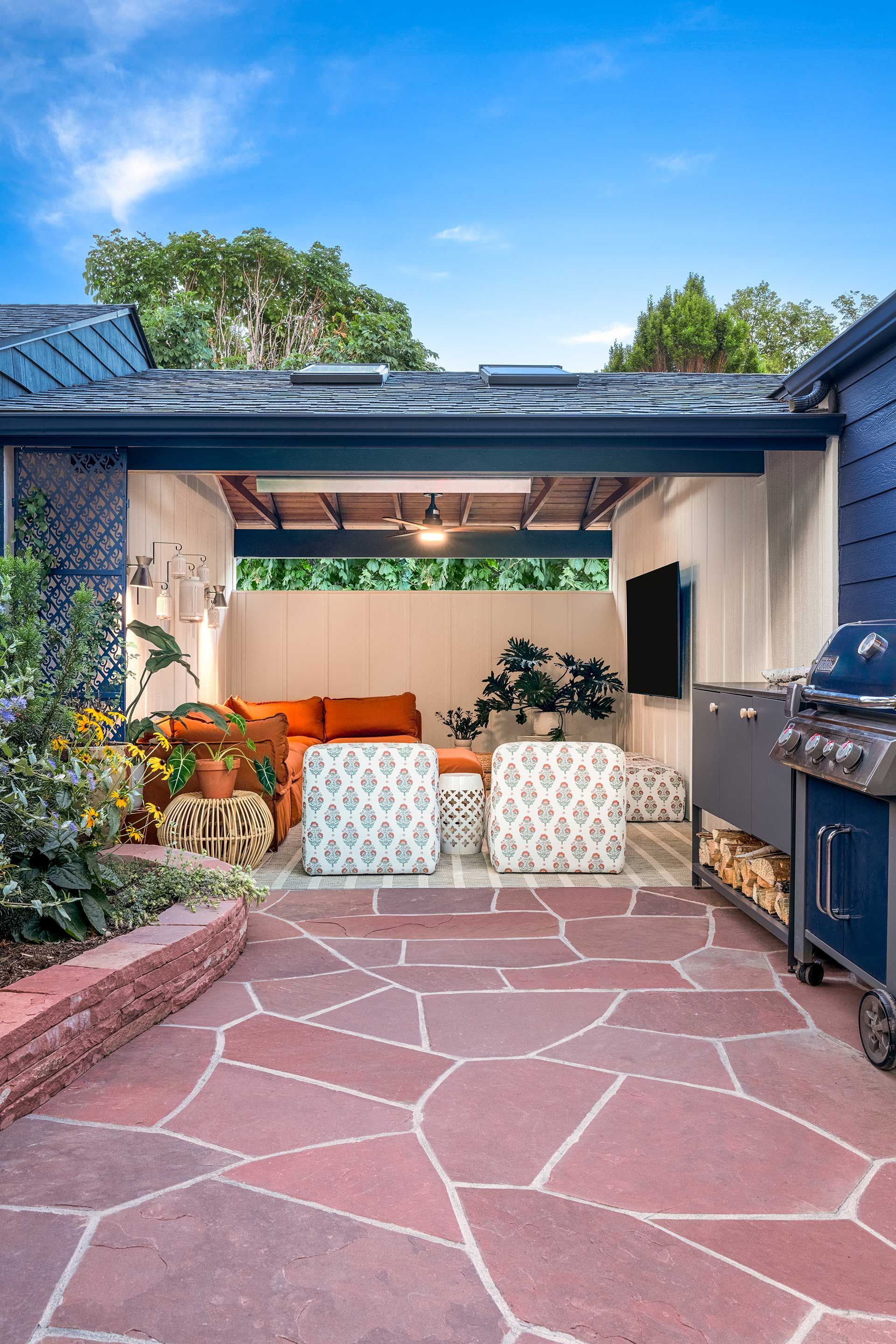 A covered outdoor patio with red stone flooring, featuring patterned armchairs, an orange sofa, and a built-in grill.