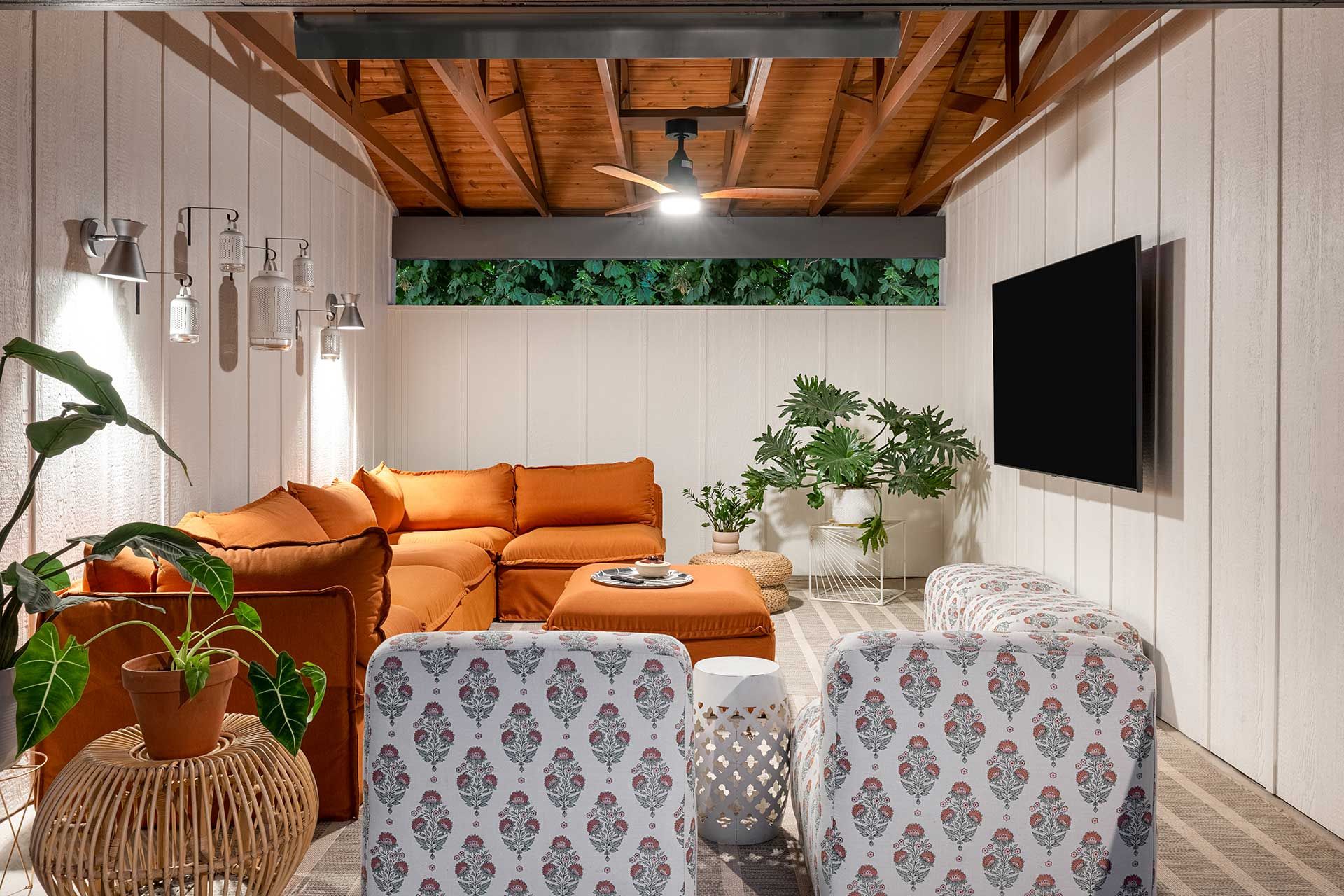 A cozy living room with a large orange sectional, patterned chairs, a wooden ceiling, and a wall-mounted television.