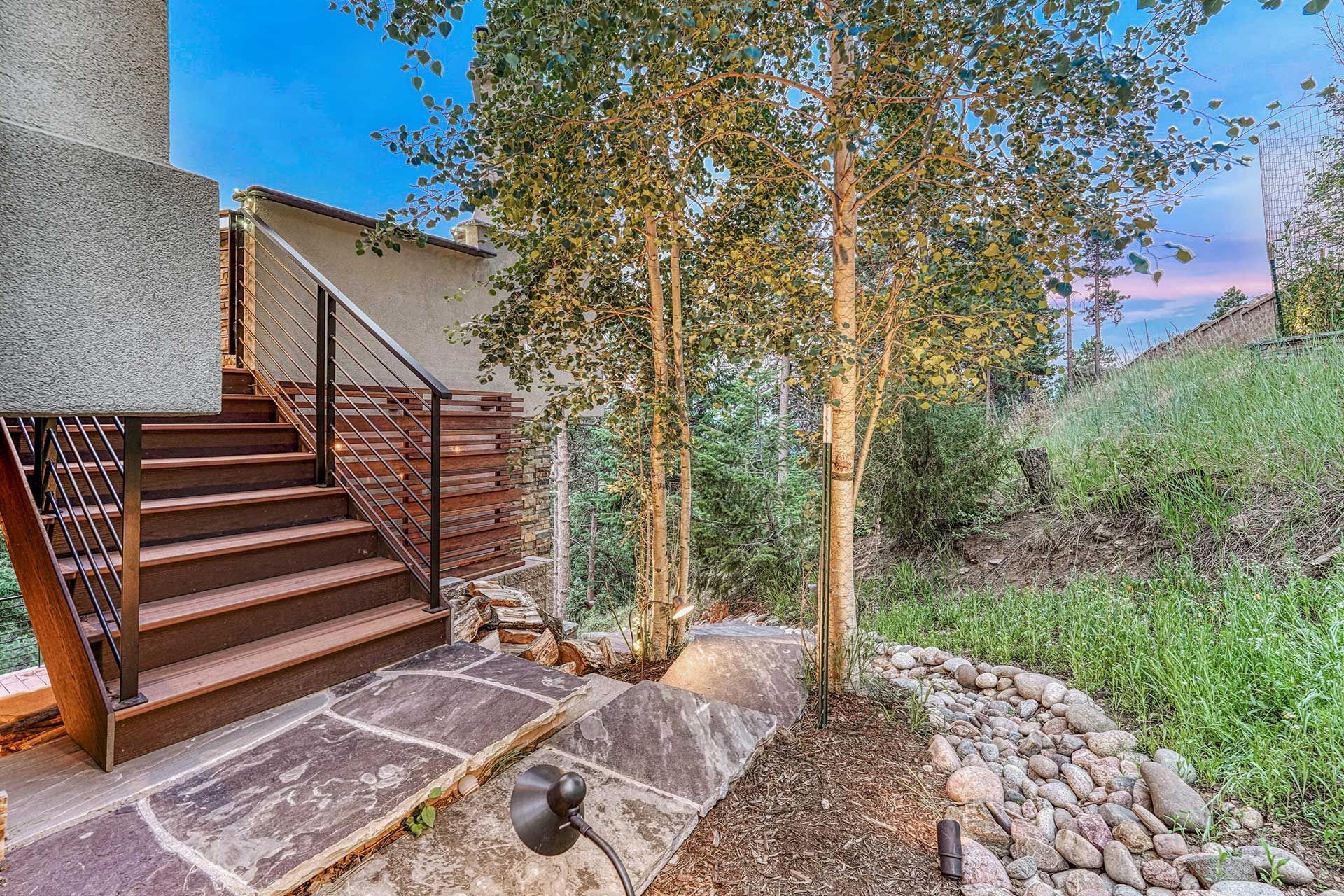 Wooden stairs lead to a stucco building beside trees and a stone pathway in a landscape at dusk.
