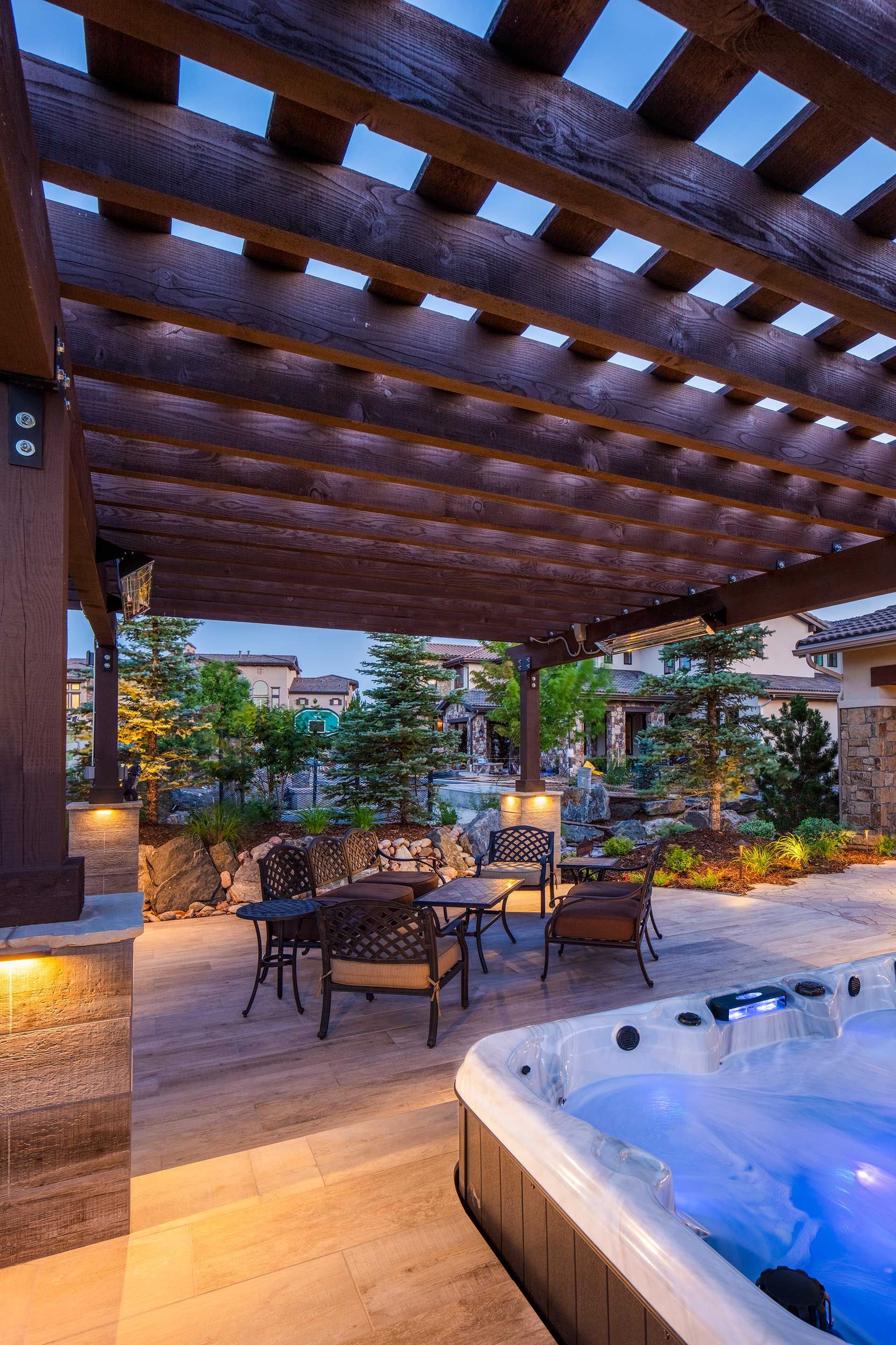A patio with a wooden pergola, outdoor seating, and a glowing hot tub at dusk, set against a landscaped garden background.