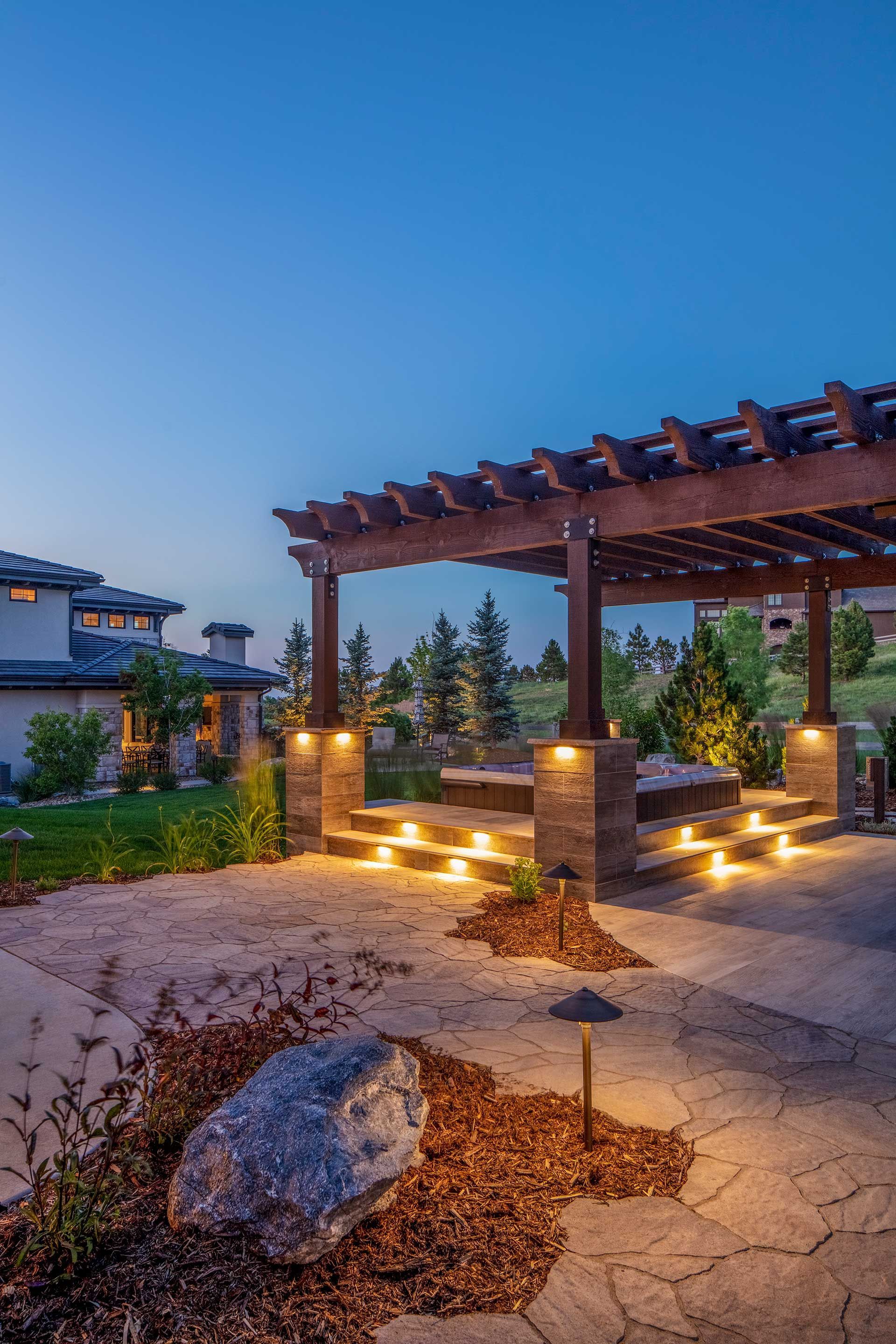 A twilight view of a landscaped backyard featuring a wooden pergola over a stone patio with warm, glowing accent lights.