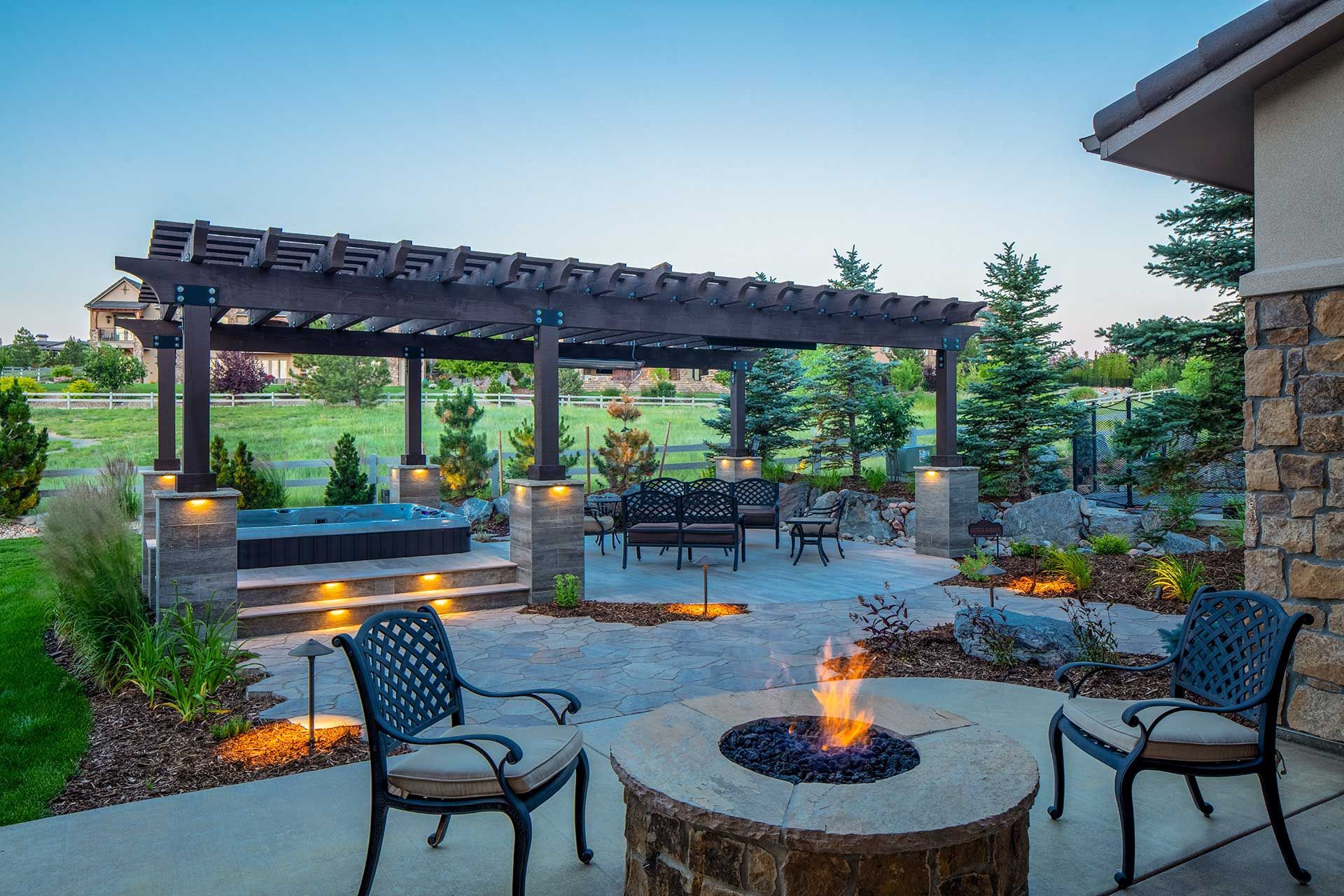 Outdoor patio with a stone fire pit, metal chairs, and a wooden pergola covering a hot tub at dusk.